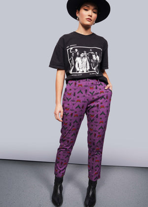 S=Person wearing black wide brimmed hat, black band t-shirt and black heeled boots has hand in pocket of purple bug printed slim crop pants.