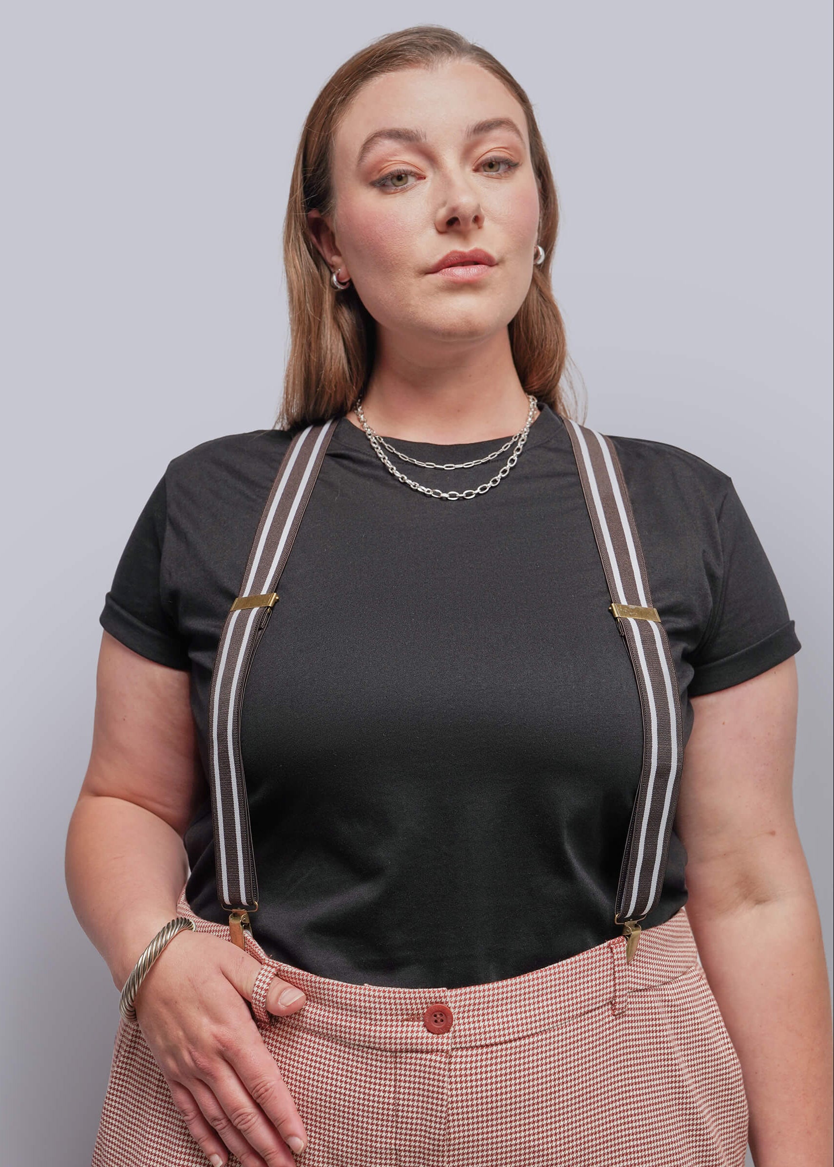 A woman modeling a black short-sleeve t-shirt with brown and cream suspenders and houndstooth trousers, set against a solid light gray studio background.