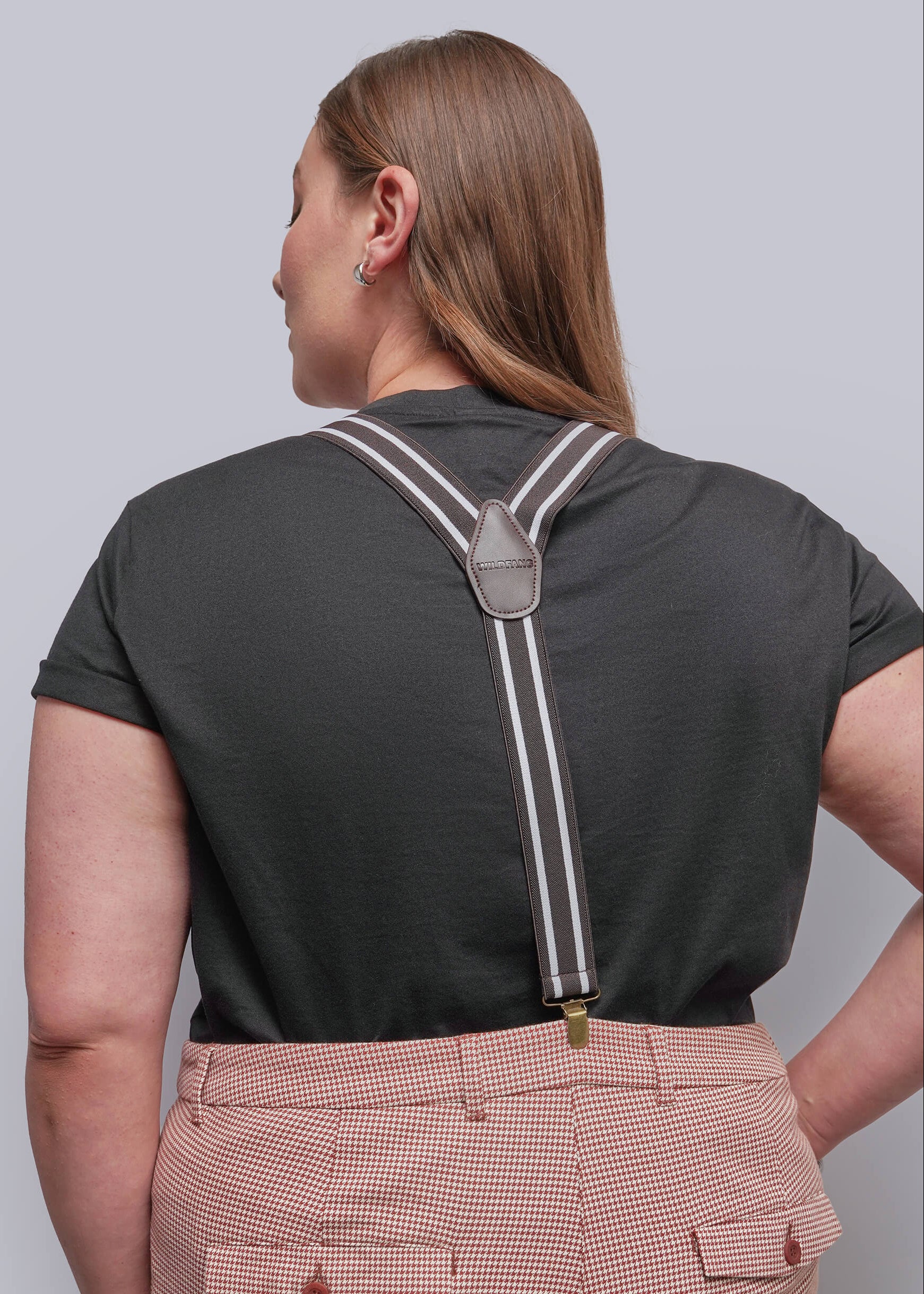 A woman shown from the back wearing brown suspenders with white stripes over a dark charcoal t-shirt and pink-and-white checkered trousers, against a solid gray studio background.