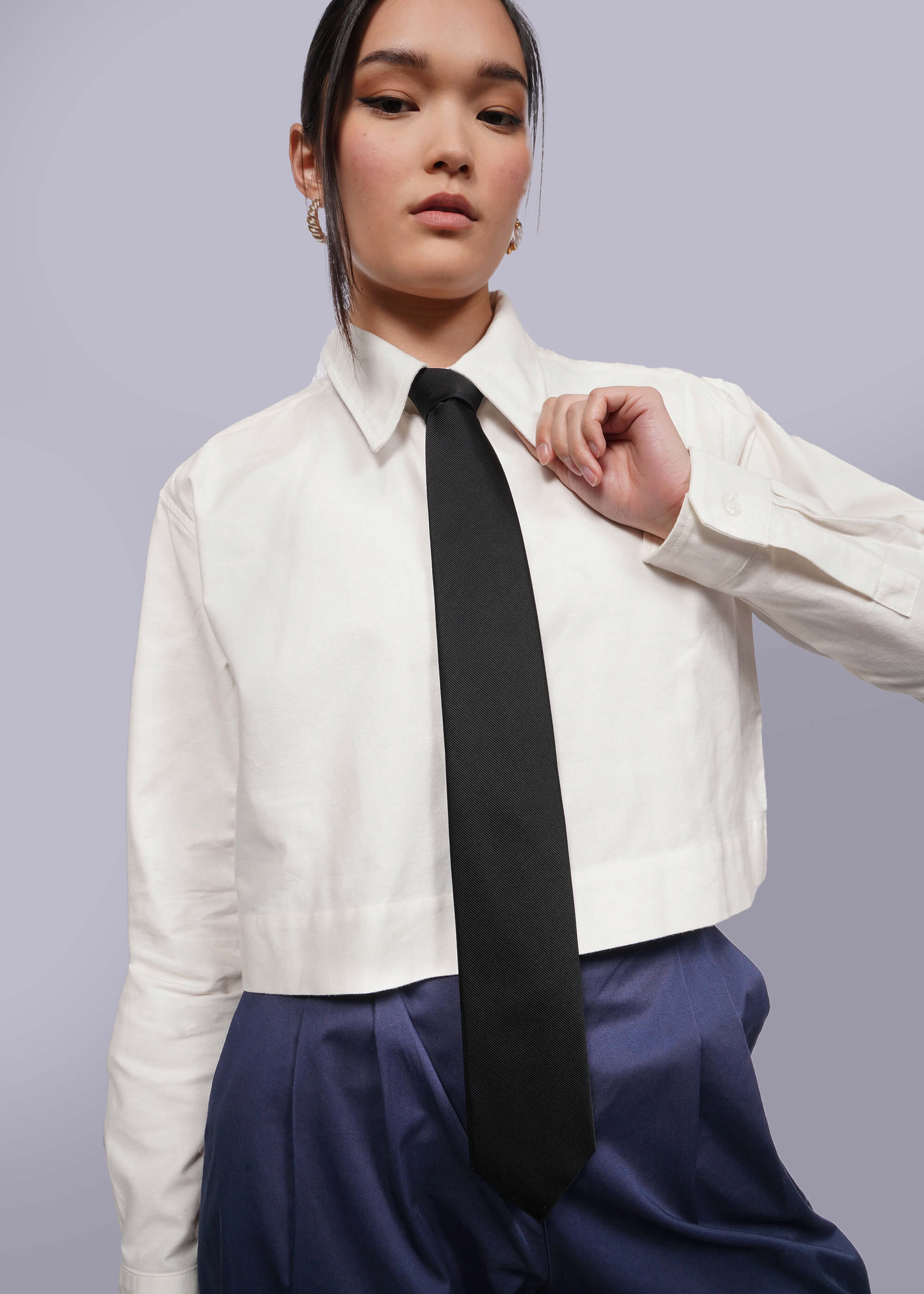 Person with black hair tied back wears cropped white button up, navy pants, and black silk tie.