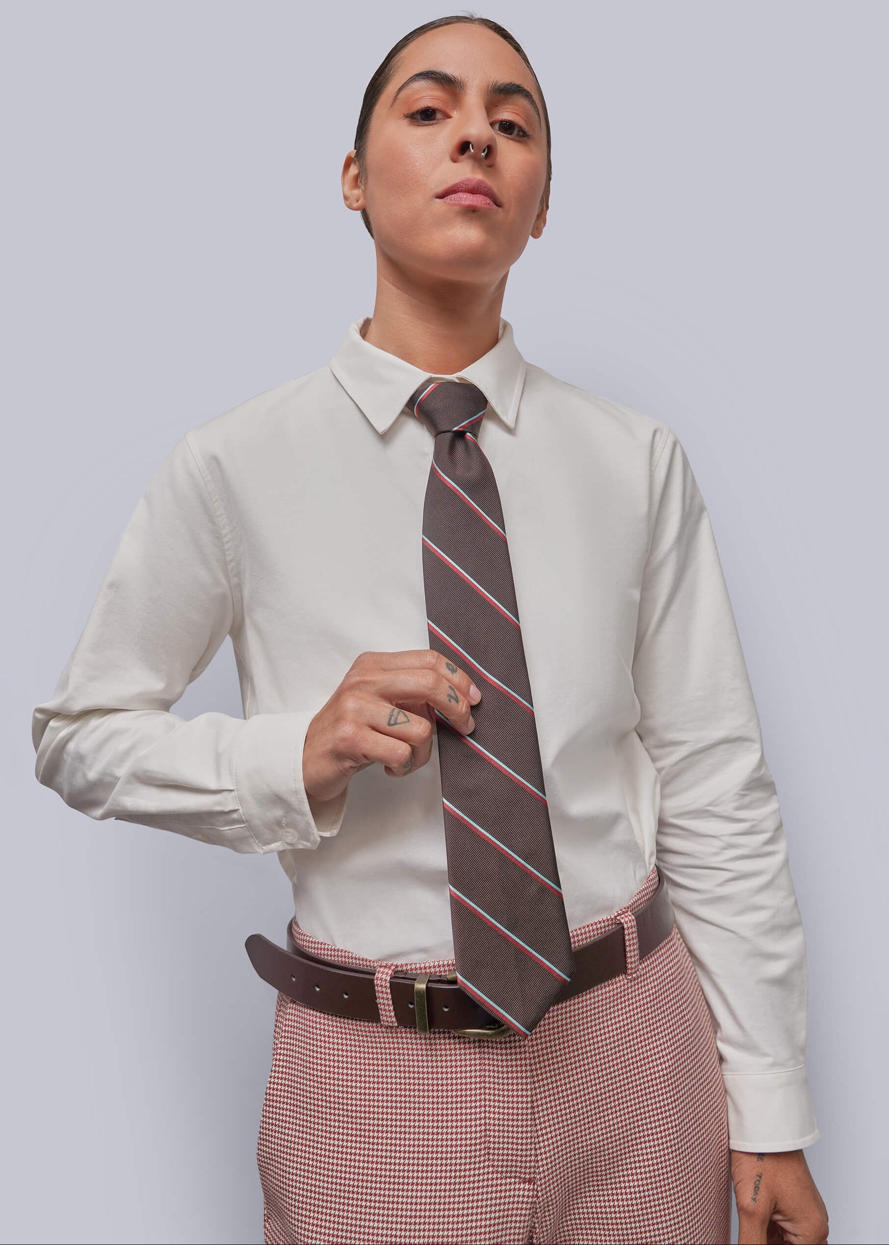 A model wearing a white long-sleeve button-down shirt, a brown striped necktie, and pink-and-white micro-check trousers, standing against a solid light gray background.