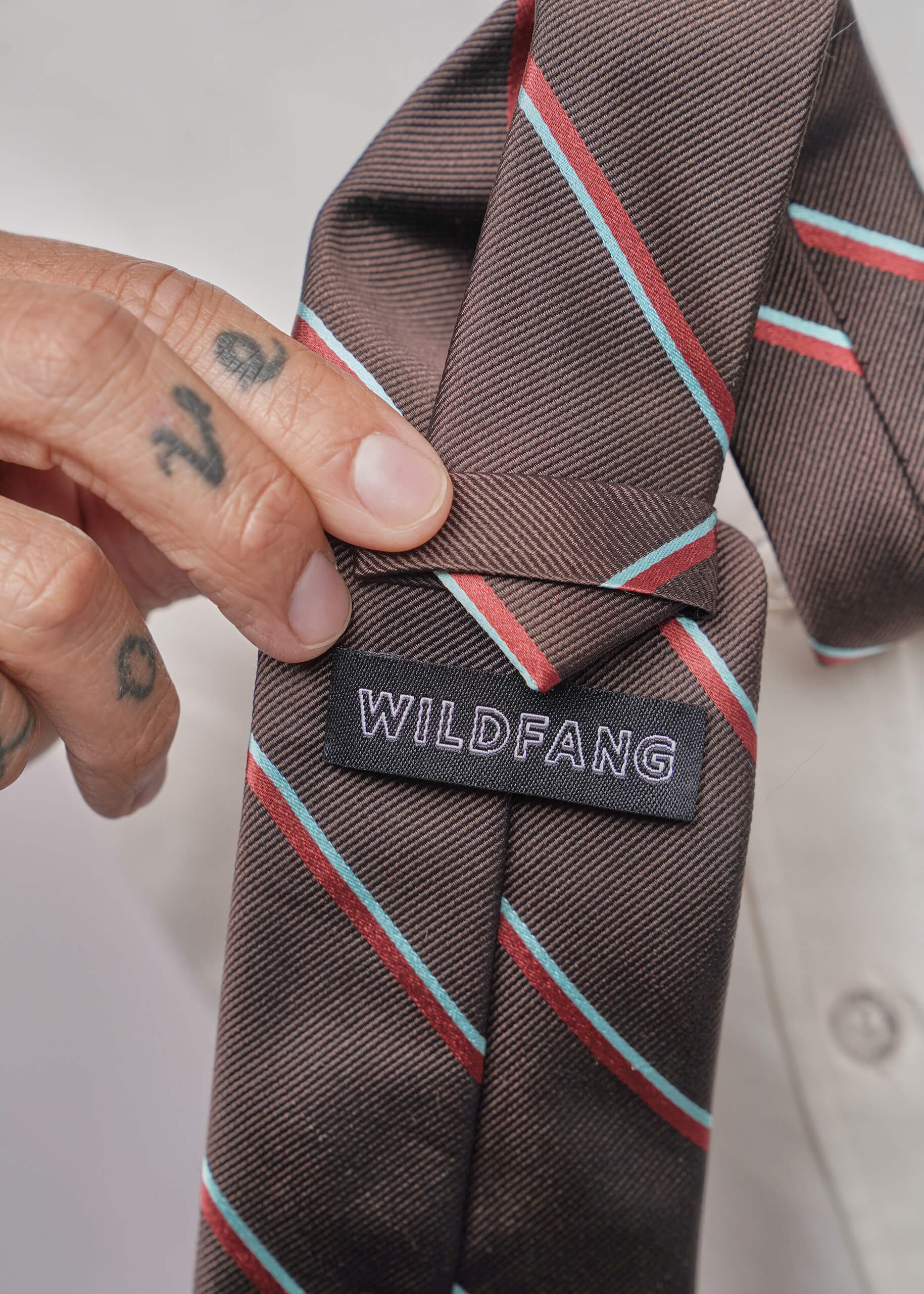 A close-up of a brown striped necktie with diagonal red and light blue lines, held by a tattooed hand. The brand label 'WILDFANG' is clearly visible on the back of the tie.
