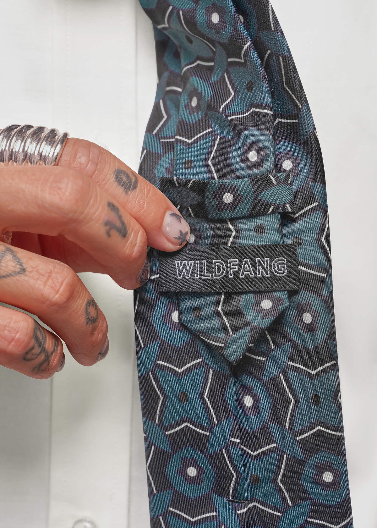 A close-up of a patterned necktie with a hand adjusting the front. The tie features a dark gray and teal geometric floral pattern with white outlines and dots.