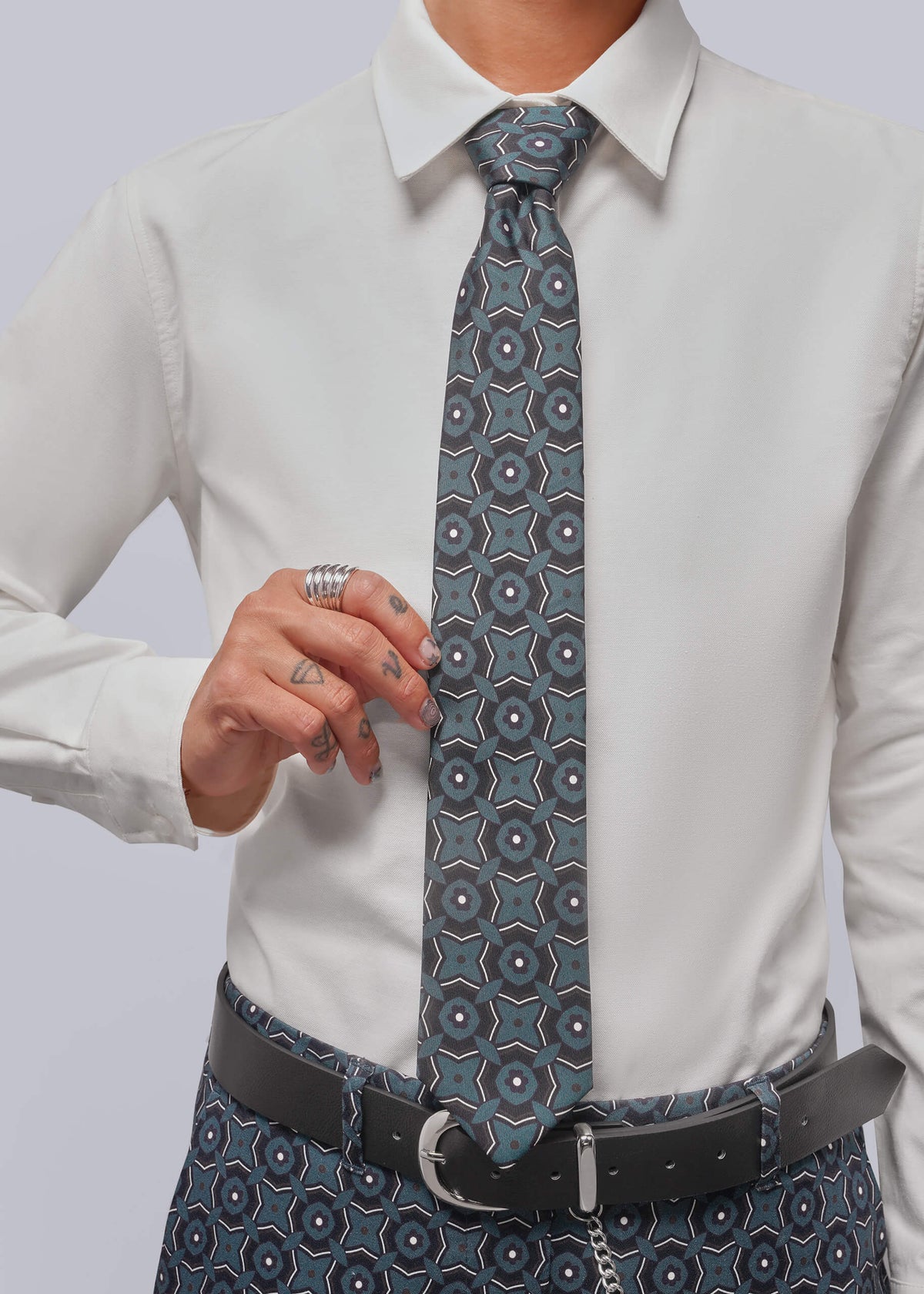 A person wearing a white dress shirt, a patterned blue-gray tie, and a black belt, shown from the chest down against a light gray studio background.