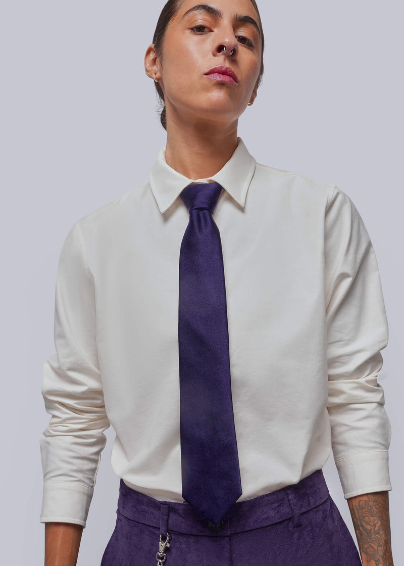 A model wearing a white long-sleeve button-down shirt, a dark purple tie, and matching purple trousers, shown from the chest up against a light gray studio background.