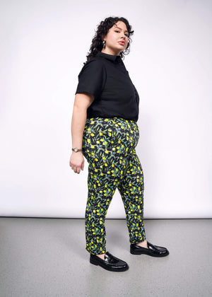 1X=Side full body view of person with curly short black hair wearing a black short sleeved button up, with The Empower Trouser in Citrus Black, which is a yellow and green print on black background featuring lemons and leaves. They also wear black loafers.