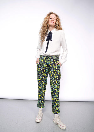 S=Front full body view of person with long curly blonde hair wearing The Empower Bow Button Up and The Empower Trouser in Citrus Black, which is a yellow and green print on black background featuring lemons and leaves. They also wear white Converse sneakers.