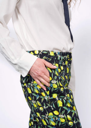 S=Close up side view of person wearing The Empower Bow Button Up and The Empower Trouser in Citrus Black, which is a yellow and green print on black background featuring lemons and leaves. They have their hand in the pant pocket.