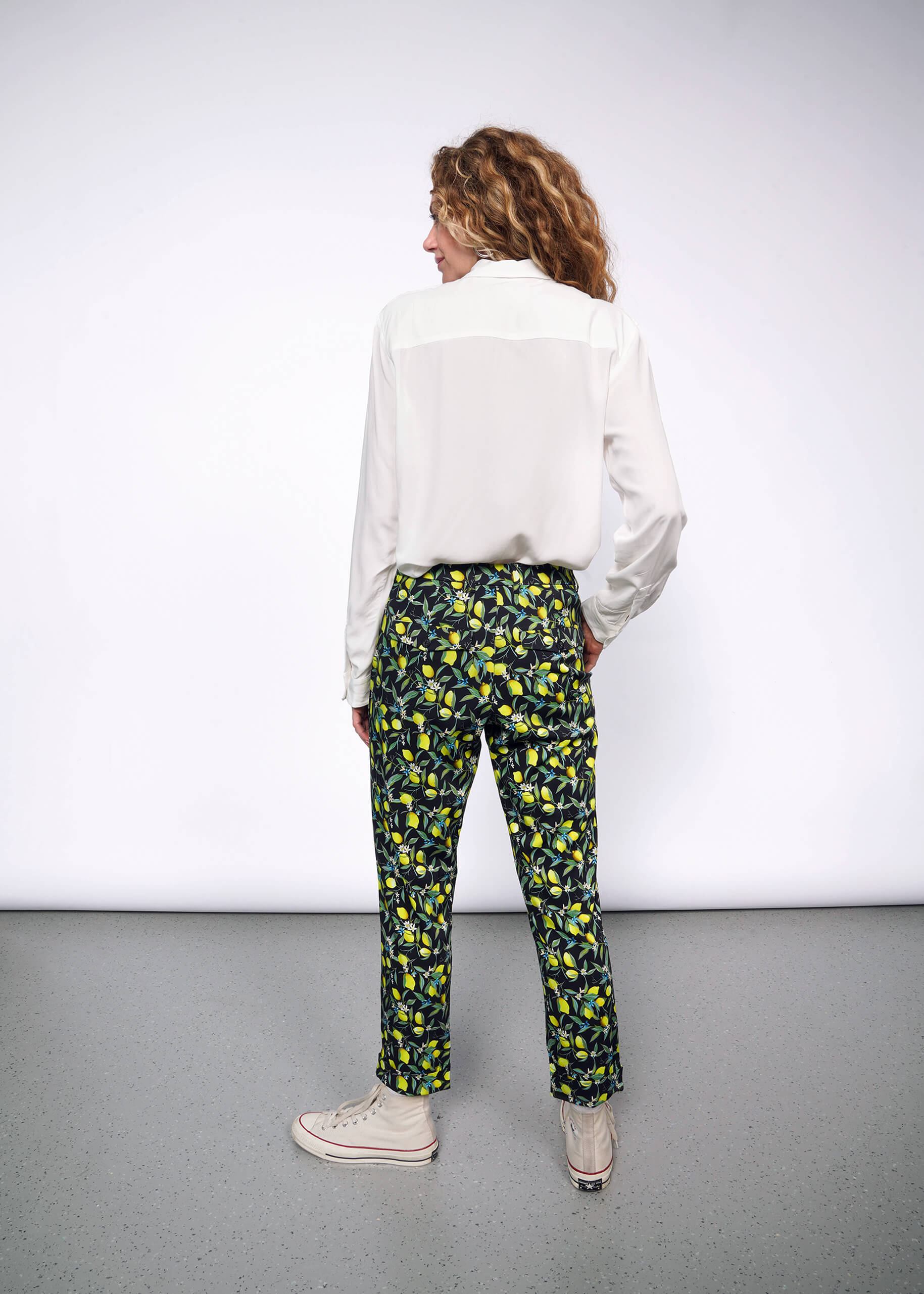 Back full body view of person with long curly blonde hair wearing The Empower Bow Button Up and The Empower Trouser in Citrus Black, which is a yellow and green print on black background featuring lemons and leaves. They also wear white Converse sneakers.