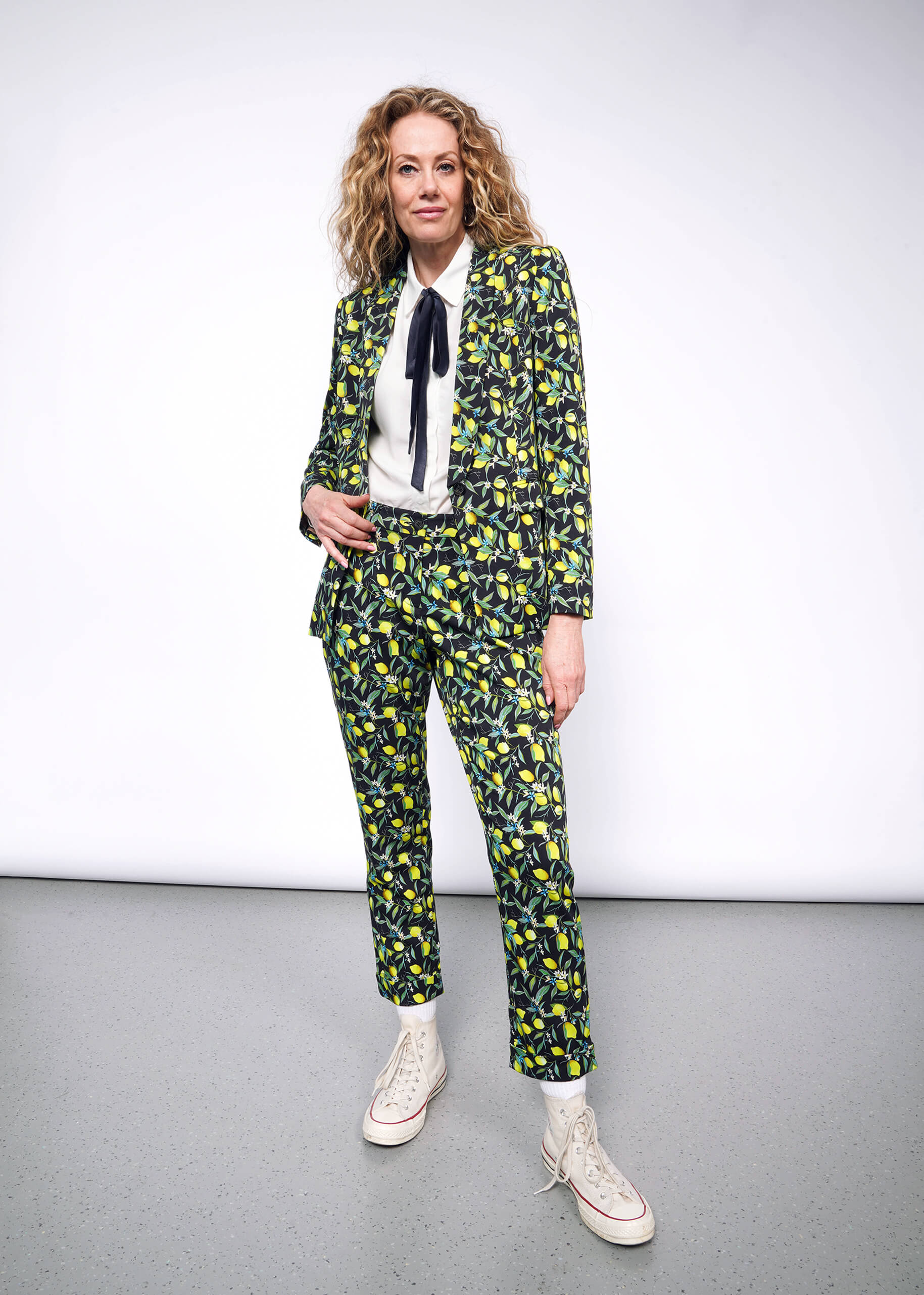 Front full body view of person with curly blonde hair wearing The Empower Bow Button Up in white, with The Empower Tux Blazer and The Empower Trouser in Citrus Black, which is a yellow and green print on black background featuring lemons and leaves. They also wear white Converse sneakers.