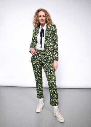 S=Front full body view of person with curly blonde hair wearing The Empower Bow Button Up in white, with The Empower Tux Blazer and The Empower Trouser in Citrus Black, which is a yellow and green print on black background featuring lemons and leaves. They also wear white Converse sneakers.