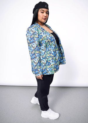 1X= Side view of person with long black hair looking at camera wearing Kangol hat, black pants, white sneakers, and open floral patterned blazer.