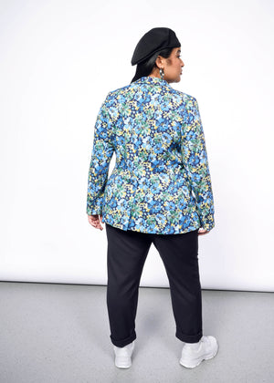 1X=Back view of person with long black hair wearing Kangol hat, black pants, white sneakers, and floral patterned blazer.