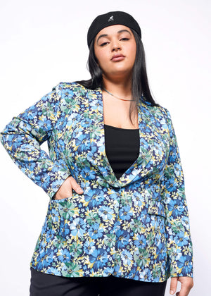 1X= Person with long black hair looks at camera wearing Kangol hat and floral patterned blazer with hand in pocket