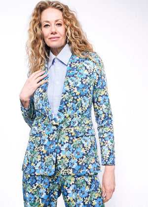 S= Person with wavy blonde hair looks at camera wearing light blue button up and floral patterned suit.