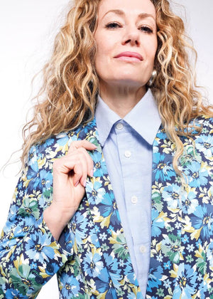 S=Close up of person with wavy blonde hair holding lapel of floral patterned blazer.
