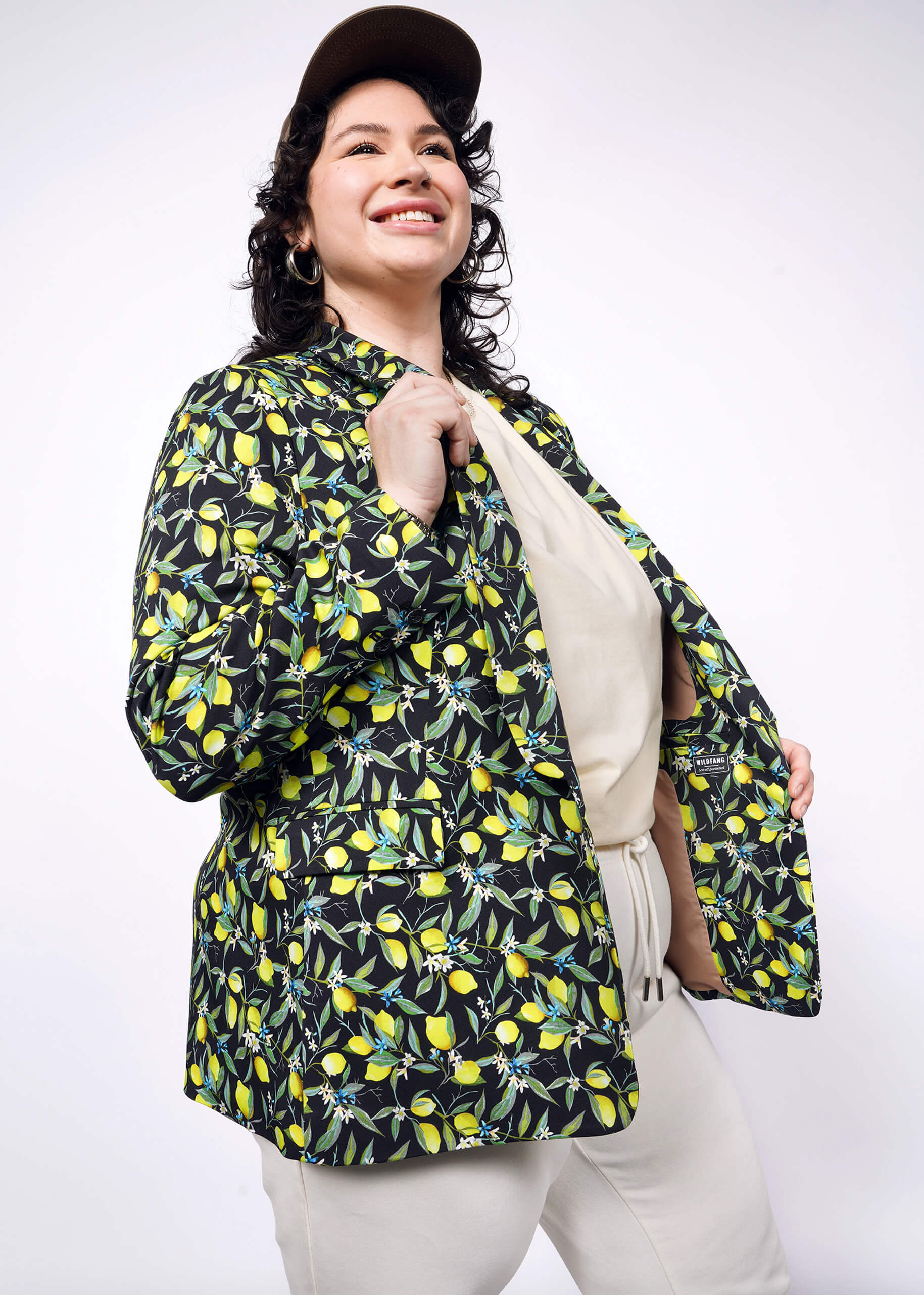 Side view of person with short curly black hair wearing black cap, tan colored shirt and pants, with The Empower Tux Blazer in Citrus Black, which is a yellow and green print on black background featuring lemons and leaves.