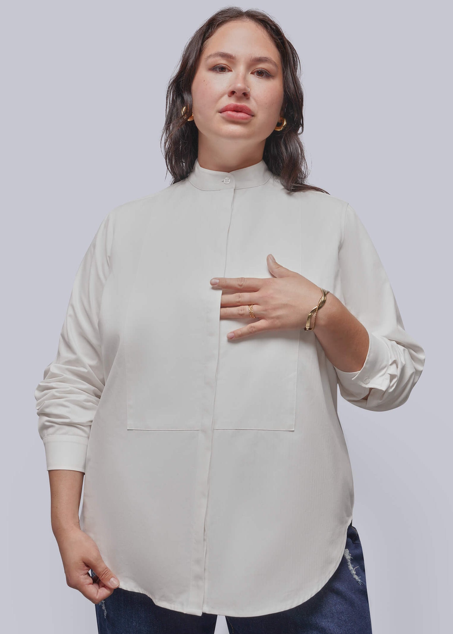 1X=A woman modeling a high-neck, long-sleeve blouse in a light cream or off-white color, paired with dark blue jeans, against a solid light gray studio background.
