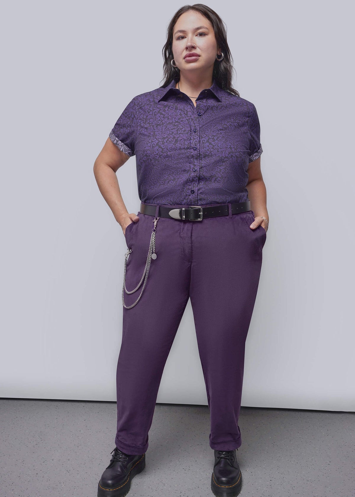 1X=A person modeling a purple short-sleeve button-up shirt with a subtle tonal pattern and matching purple trousers, styled with a black belt, silver chain detail, and black boots against a plain studio background.