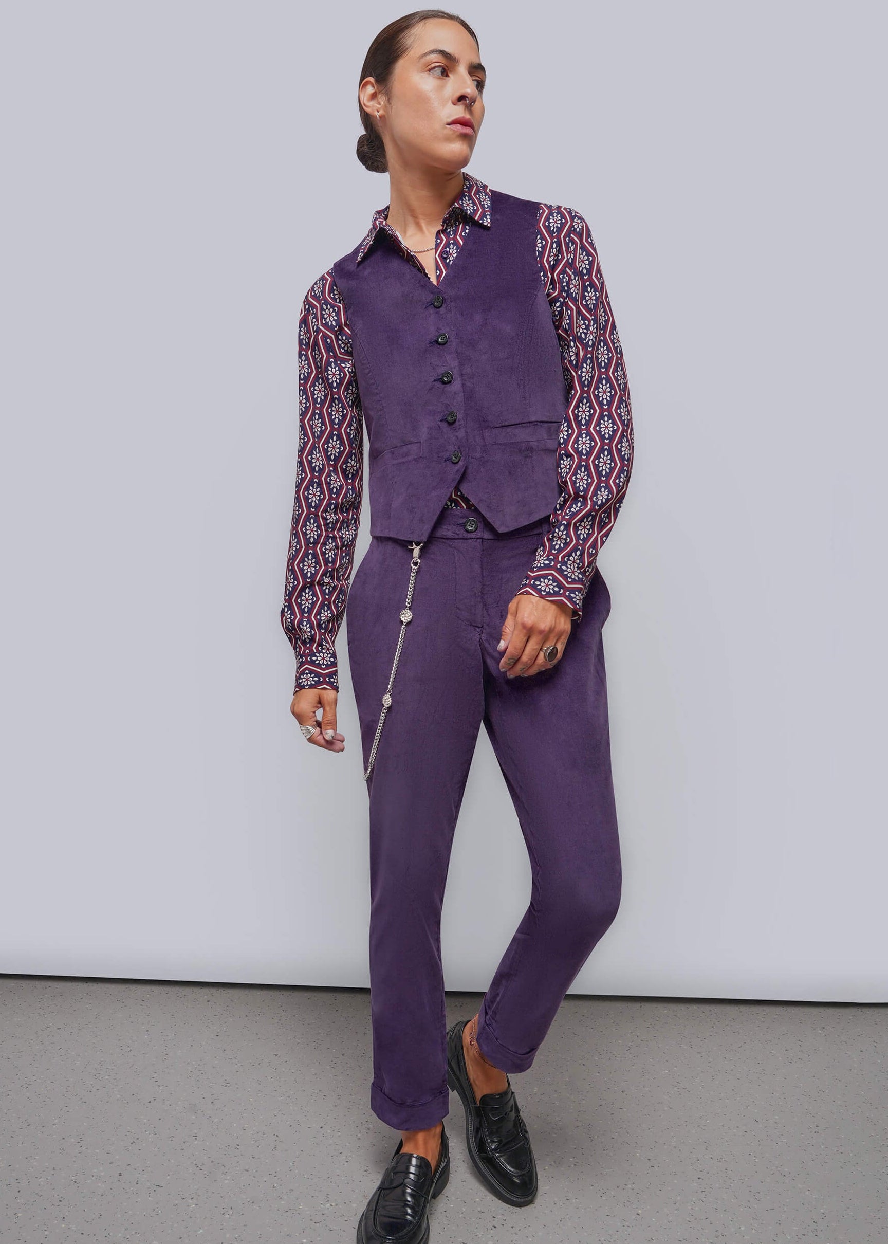 A model wearing a purple vest and matching trousers with a patterned long-sleeve shirt and black leather shoes, standing against a light gray studio background.