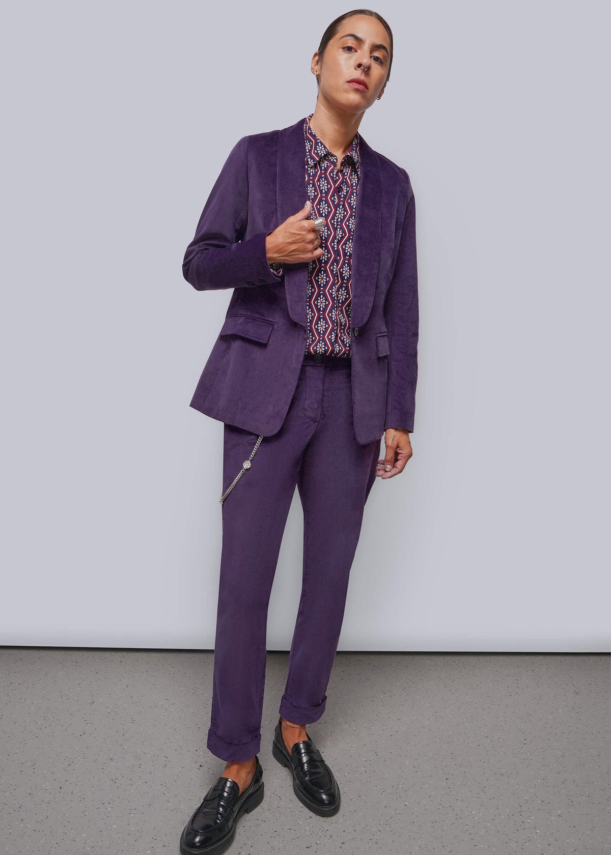 S=A model wearing a purple velvet blazer and matching trousers with a patterned shirt and black loafers, standing against a light gray studio background.