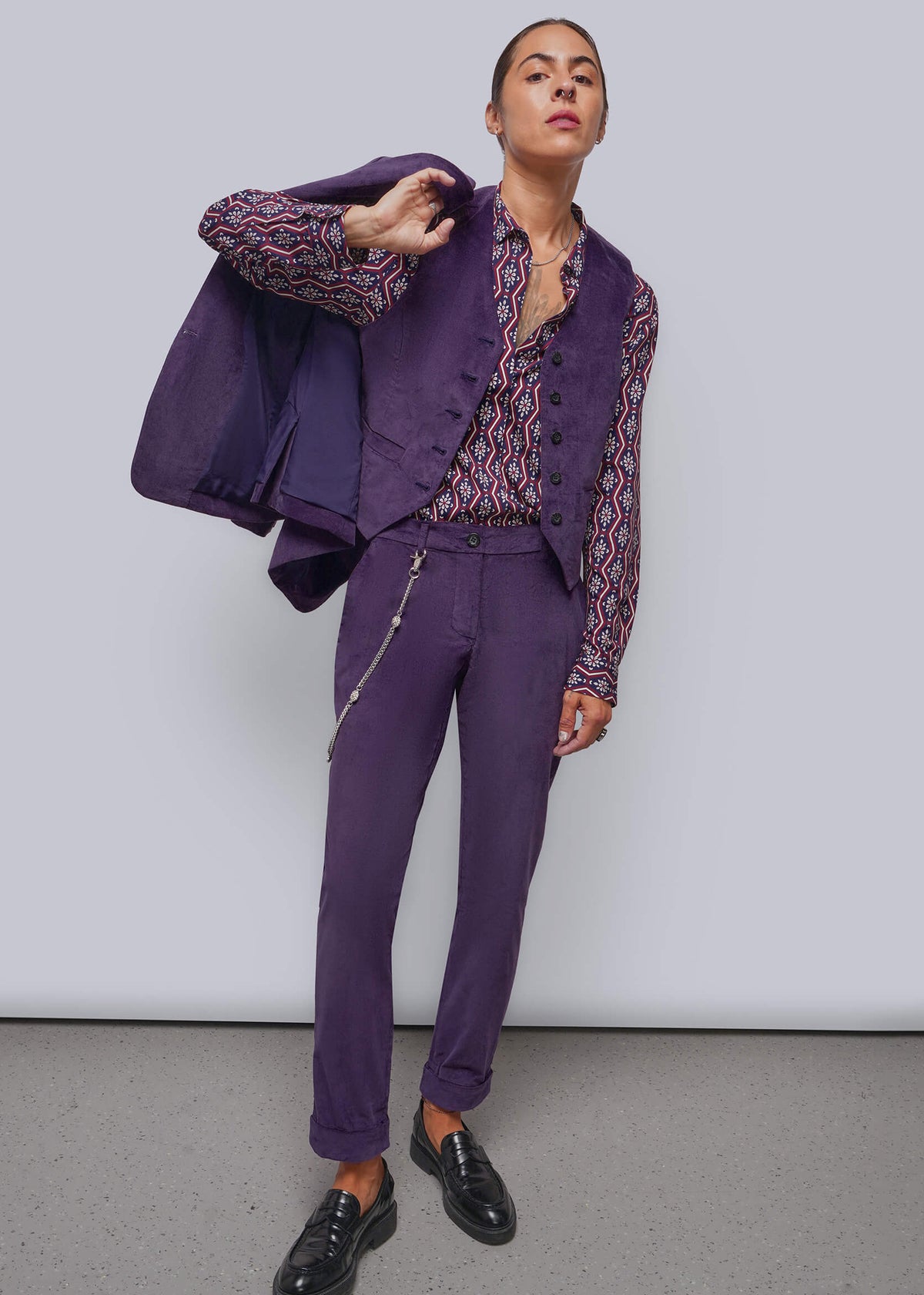 S=A model wearing a purple tailored suit consisting of a vest, patterned shirt, and trousers, with a matching purple blazer draped over one shoulder, set against a plain light gray studio background.