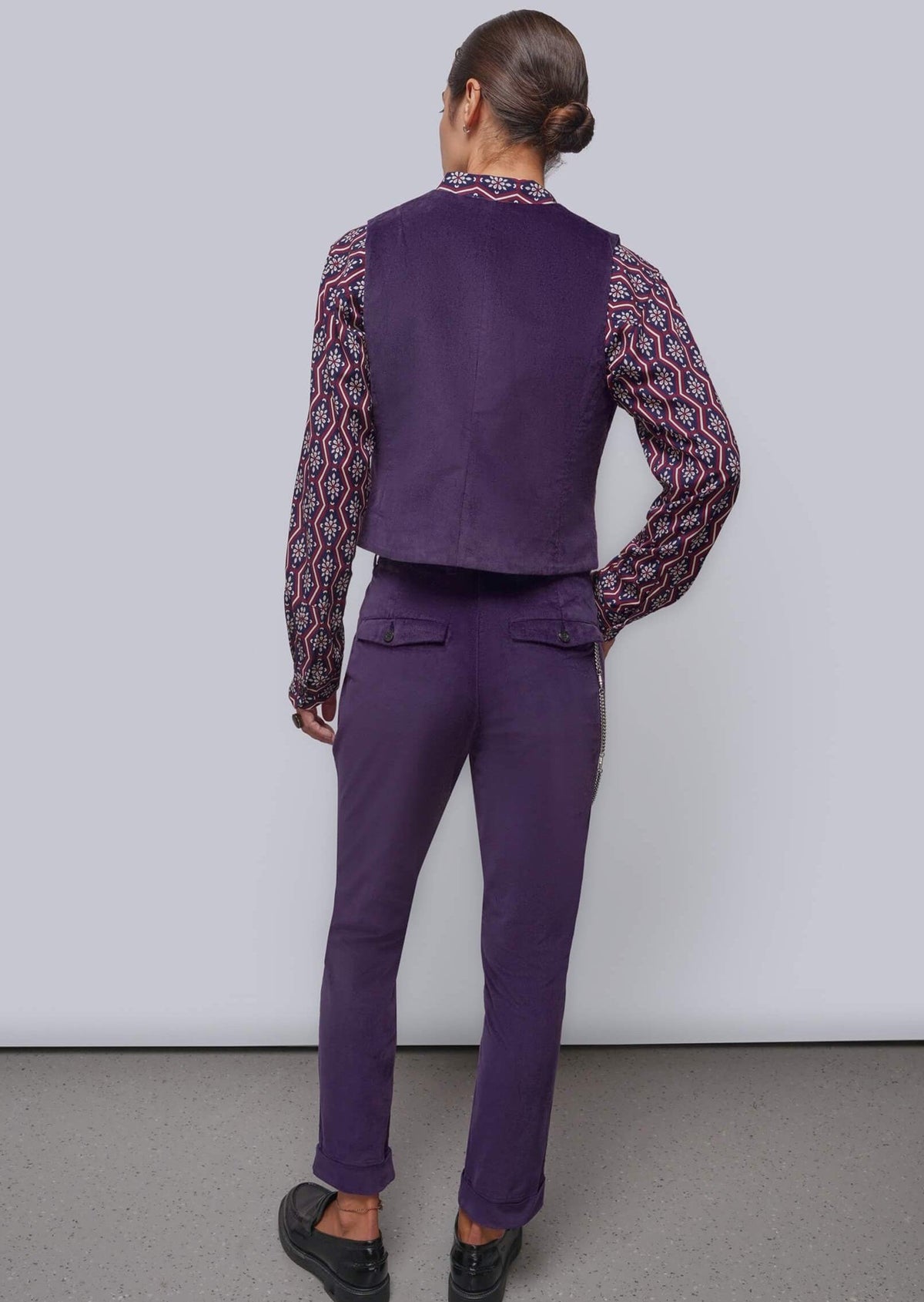 S=A model shown from the back wearing a matching purple vest and trousers set, layered over a long-sleeve shirt with a red, black, and white geometric pattern, paired with black shoes, against a plain light gray studio background.