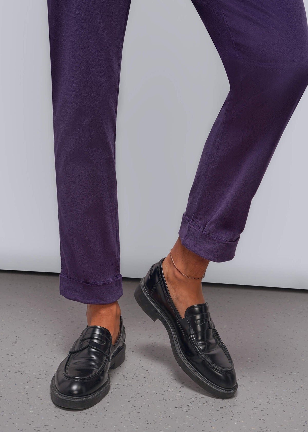 S=A person wearing purple trousers and black leather penny loafers, shown from the knees down against a light gray studio background.