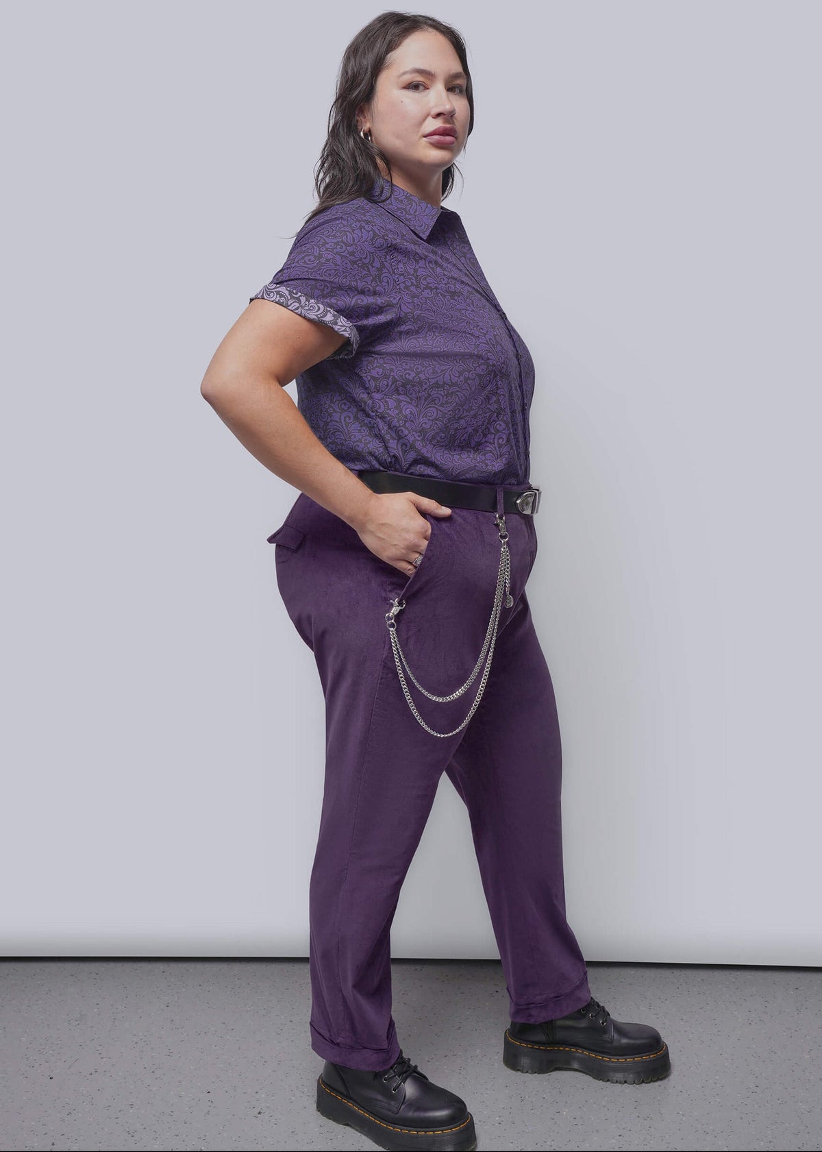 1X=A woman modeling a purple short-sleeve patterned button-up shirt and matching purple trousers, styled with a black belt, silver chain accessory, and black platform shoes, against a light gray studio background.