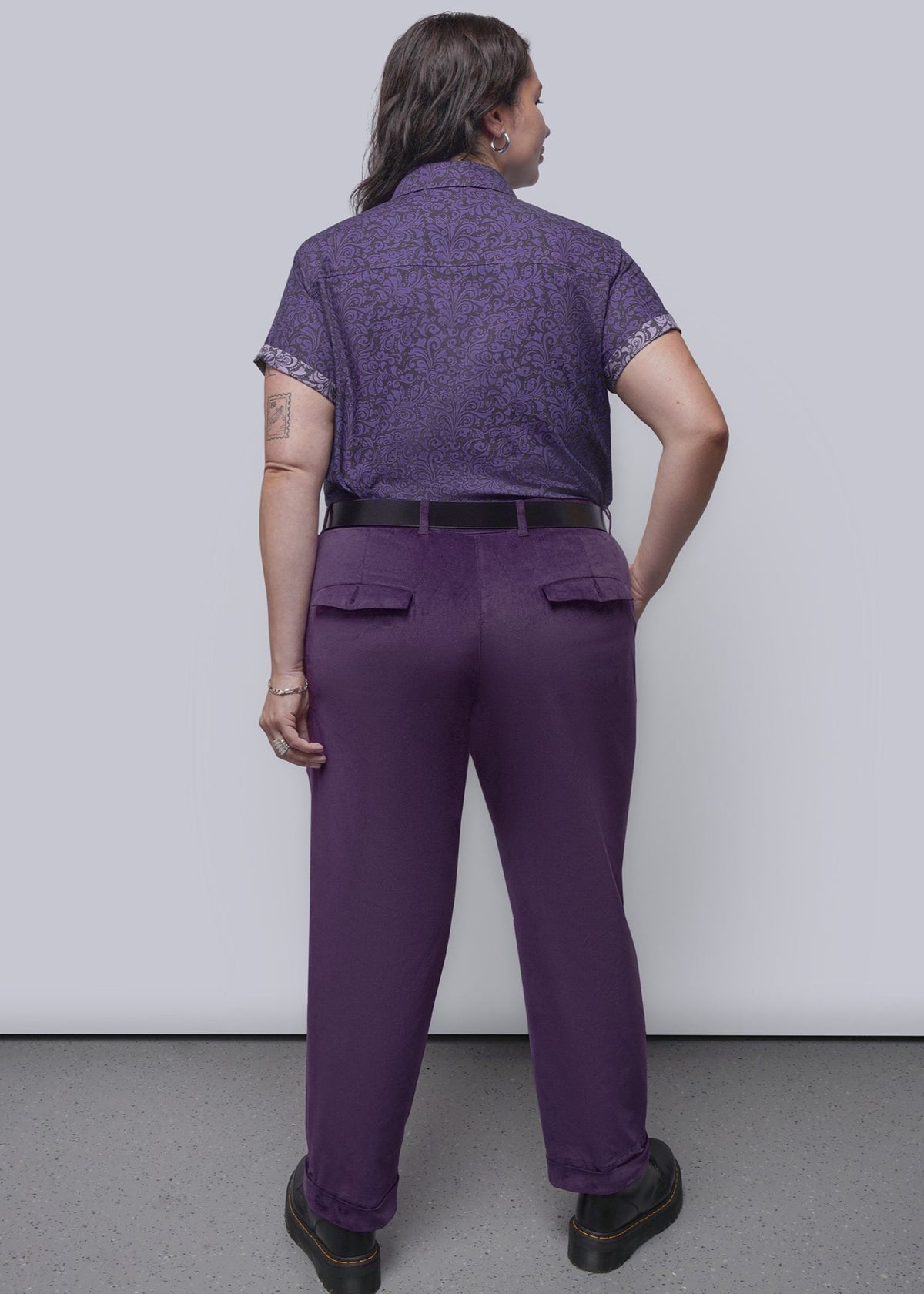1X=A person shown from the back wearing a purple short-sleeve patterned shirt and matching purple trousers, standing against a plain light gray wall and gray floor.