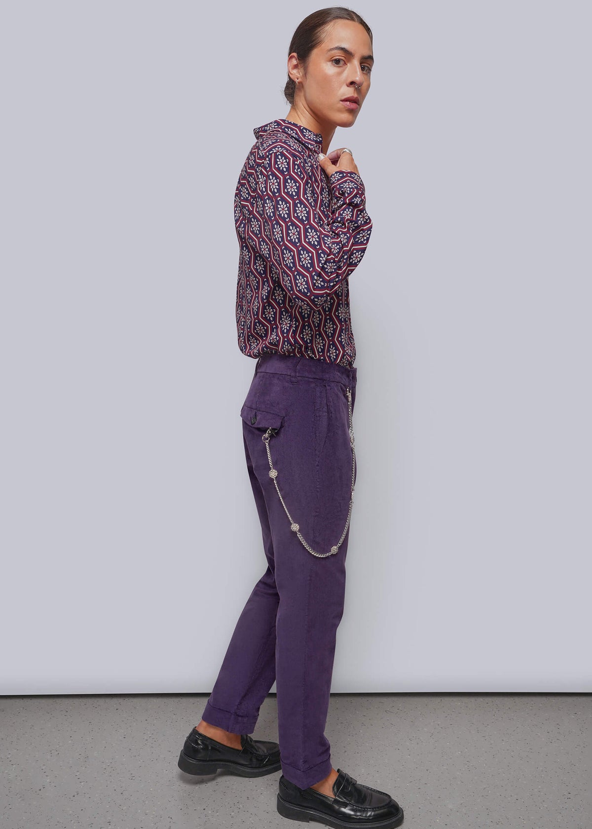 S=A model wearing purple velvet pants with a silver chain detail, paired with a multicolor patterned long-sleeve shirt and black leather loafers, standing against a light gray studio background.