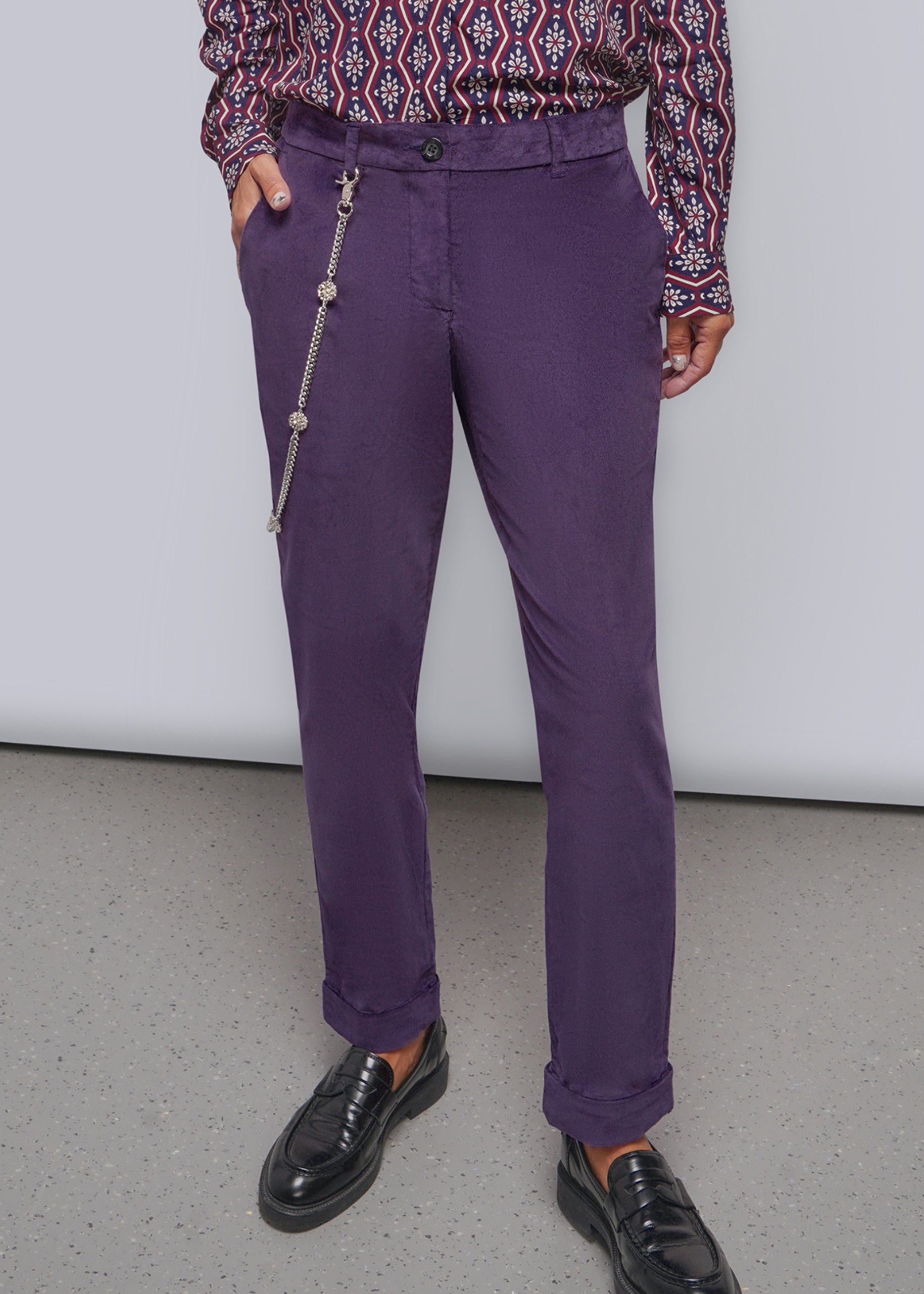 A person wearing purple trousers with a silver wallet chain hanging from the side pocket, paired with a patterned long-sleeve shirt and black loafers, standing against a plain light gray wall.