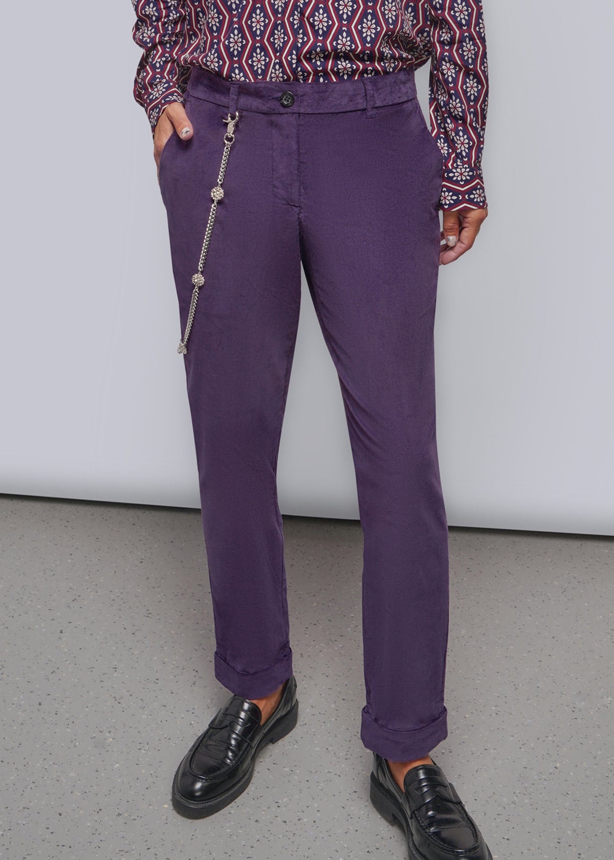 S=A person wearing purple trousers with a silver wallet chain hanging from the side pocket, paired with a patterned long-sleeve shirt and black loafers, standing against a plain light gray wall.