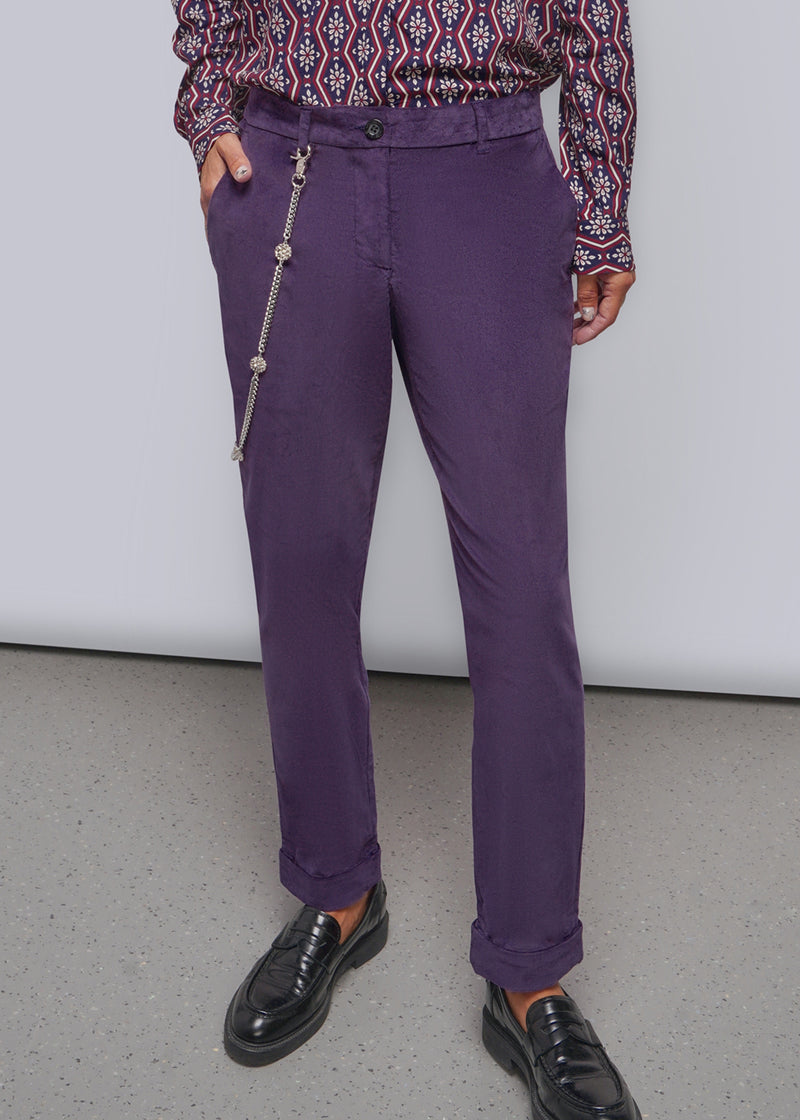 S=A person wearing purple trousers with a silver wallet chain hanging from the side pocket, paired with a patterned long-sleeve shirt and black loafers, standing against a plain light gray wall.