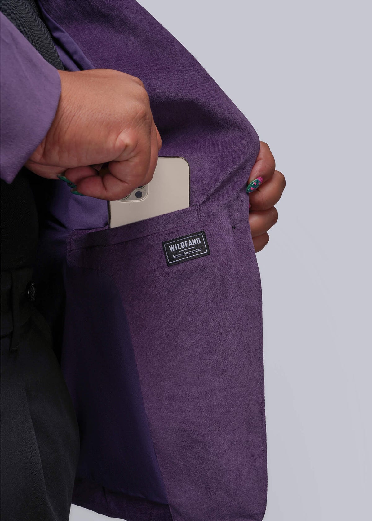 1X=A close-up of a person wearing a purple velvet blazer, showing a hand sliding a white smartphone into the side pocket. The blazer features a black brand label that reads 'WILDFANG'.