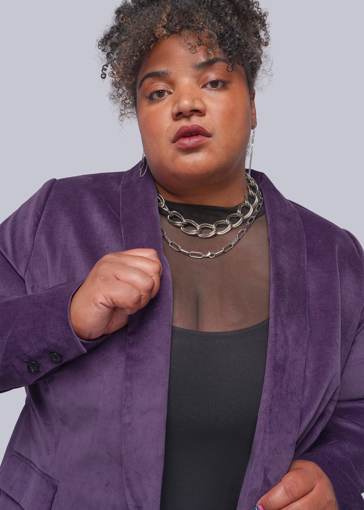 1X=A plus-size model wearing a purple velvet blazer over a black mesh top and silver chain necklaces, set against a solid gray studio background.