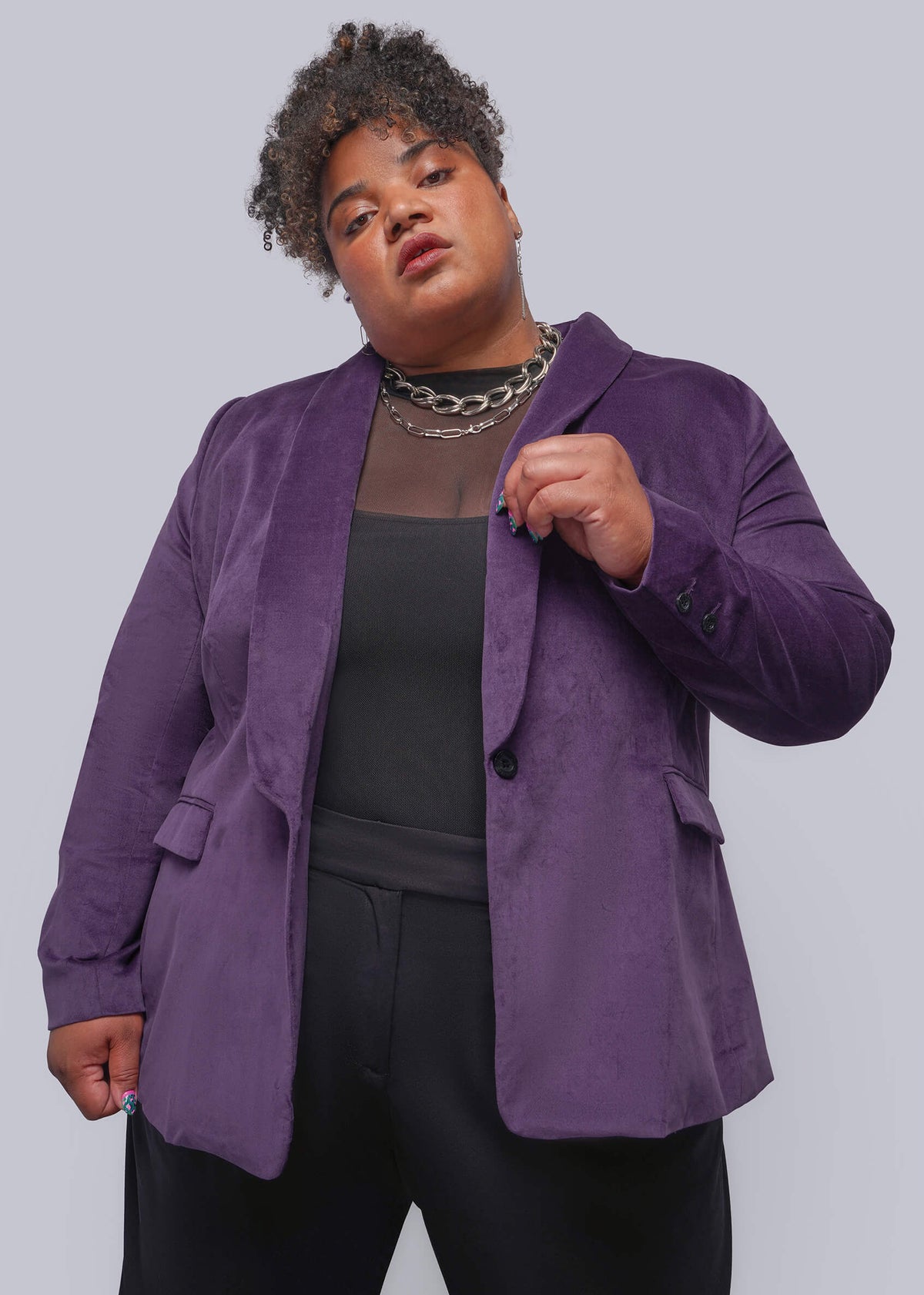 1X=A plus-size model wearing a purple velvet blazer over a black top and black pants, accessorized with a chunky silver chain necklace, against a solid gray studio background.