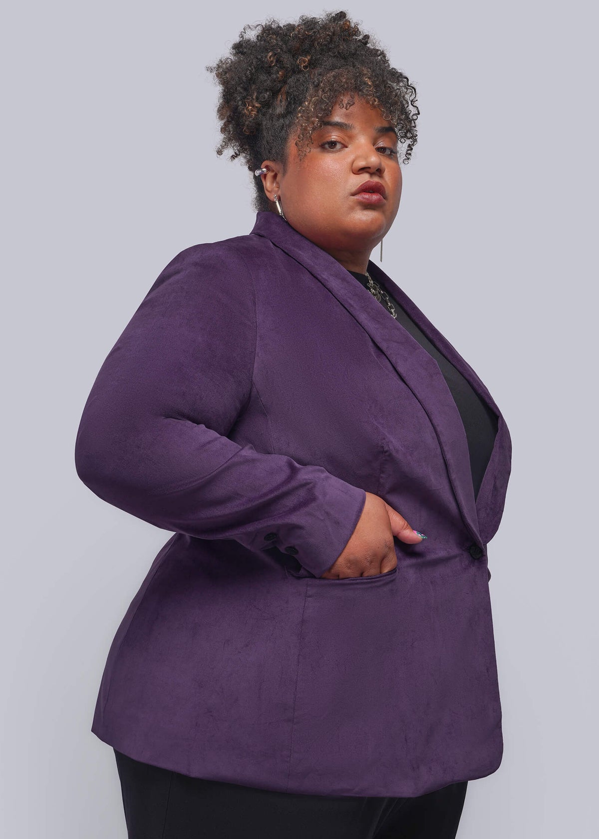 1X=A plus-size model wearing a purple velvet blazer over a black top and black pants, standing against a solid gray studio background.