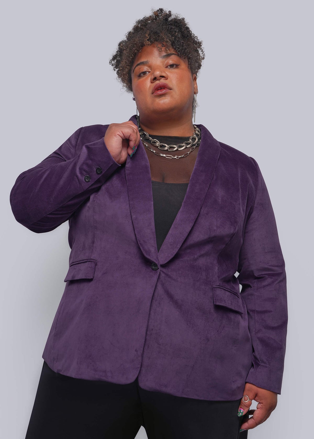1X=A plus-size model wearing a purple velvet blazer with black trousers and layered silver necklaces, posing against a solid light gray studio background.