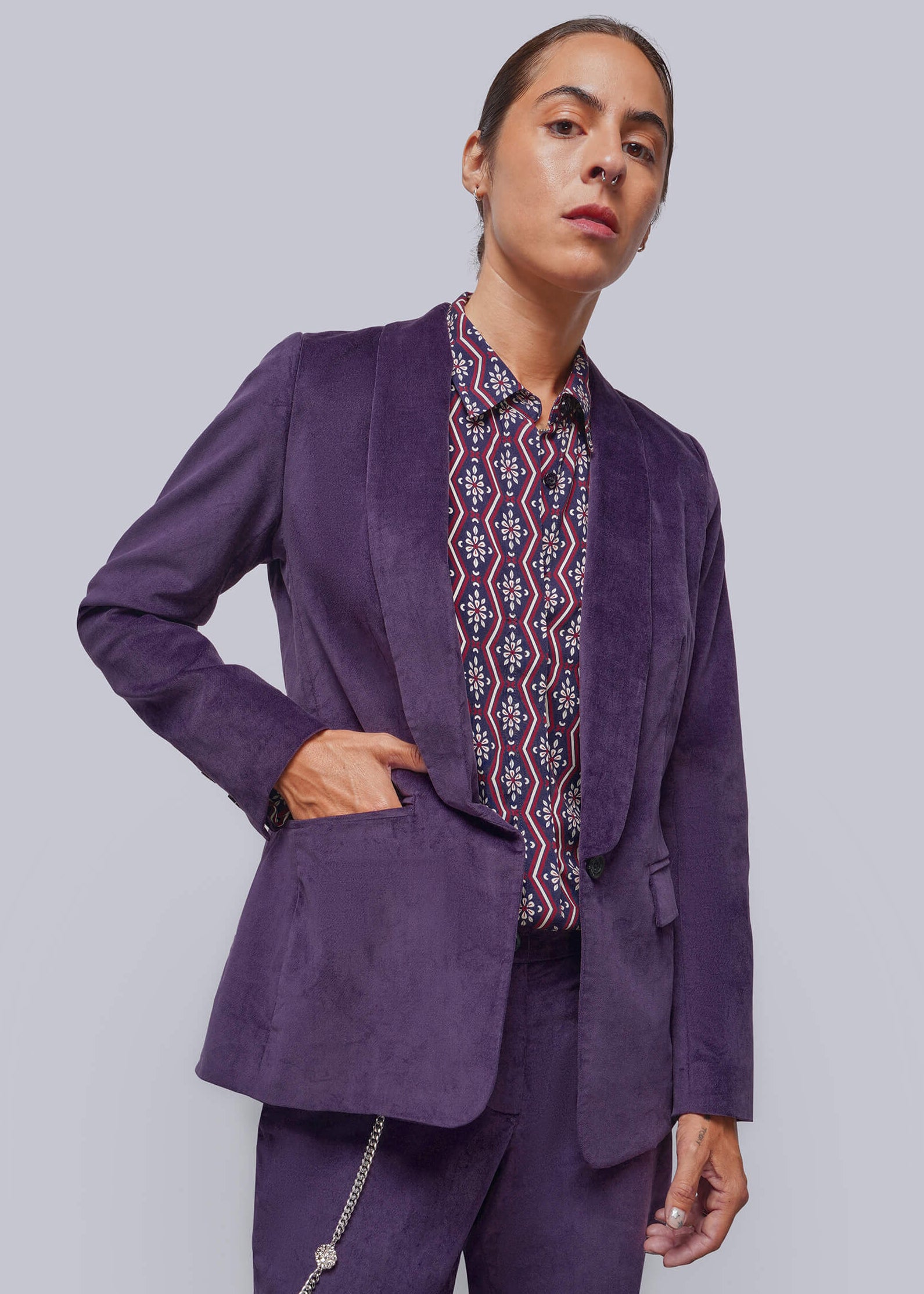 A model wearing a purple velvet blazer and matching trousers, styled with a patterned button-down shirt, against a solid gray studio background.