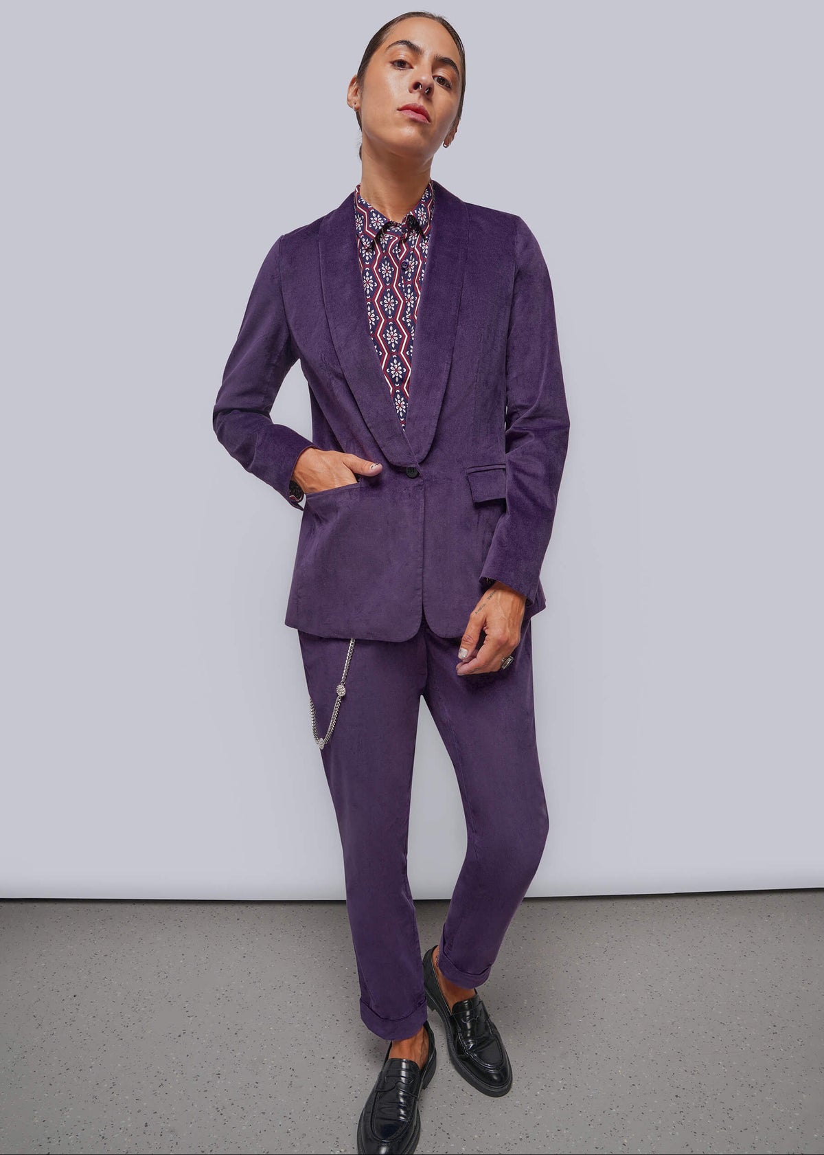 S=A model wearing a matching purple velvet blazer and trousers suit, styled with a patterned button-down shirt and black leather shoes, standing against a light gray studio background.