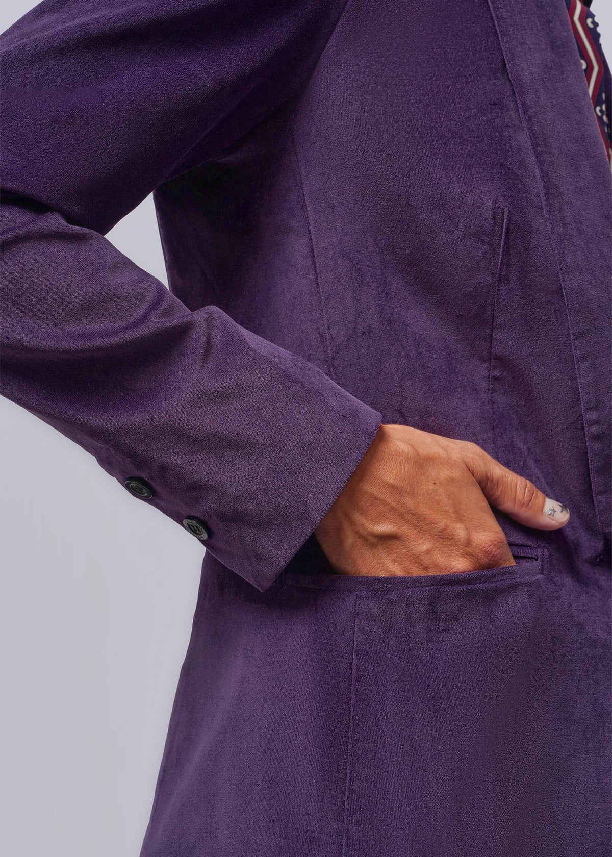 S=Close-up of a person wearing a textured purple blazer, with one hand tucked into a front pocket. The fabric has a velvet texture, and a patterned shirt is partially visible at the collar against a light gray background.