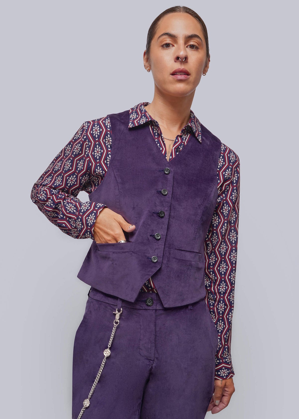 S=A model wearing a purple velvet vest and matching trousers over a patterned long-sleeve shirt, posed against a solid gray studio background.