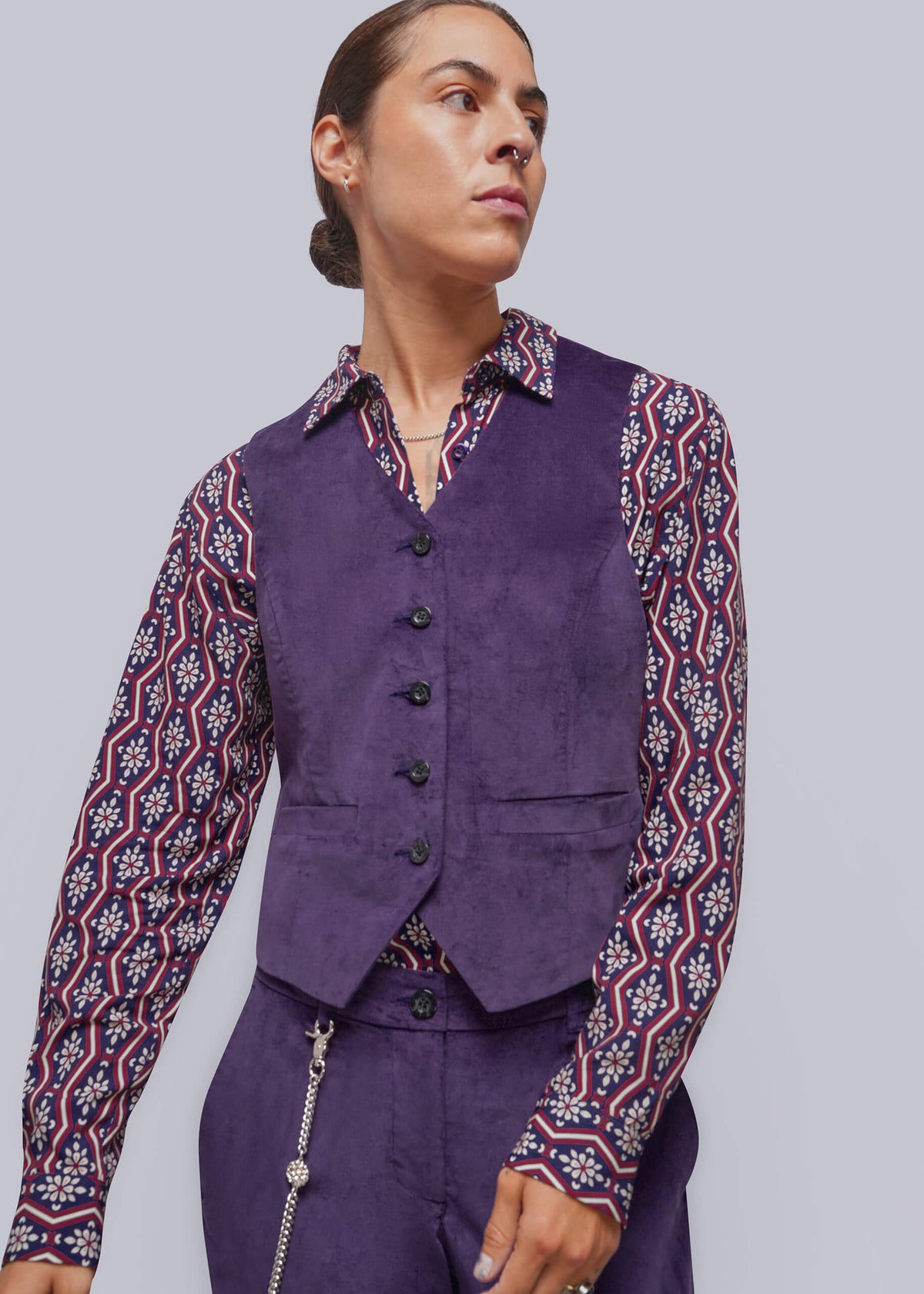 S=A model wearing a purple corduroy vest over a long-sleeve button-up shirt with a red, white, and navy geometric pattern, paired with matching purple trousers, shot against a solid gray background.