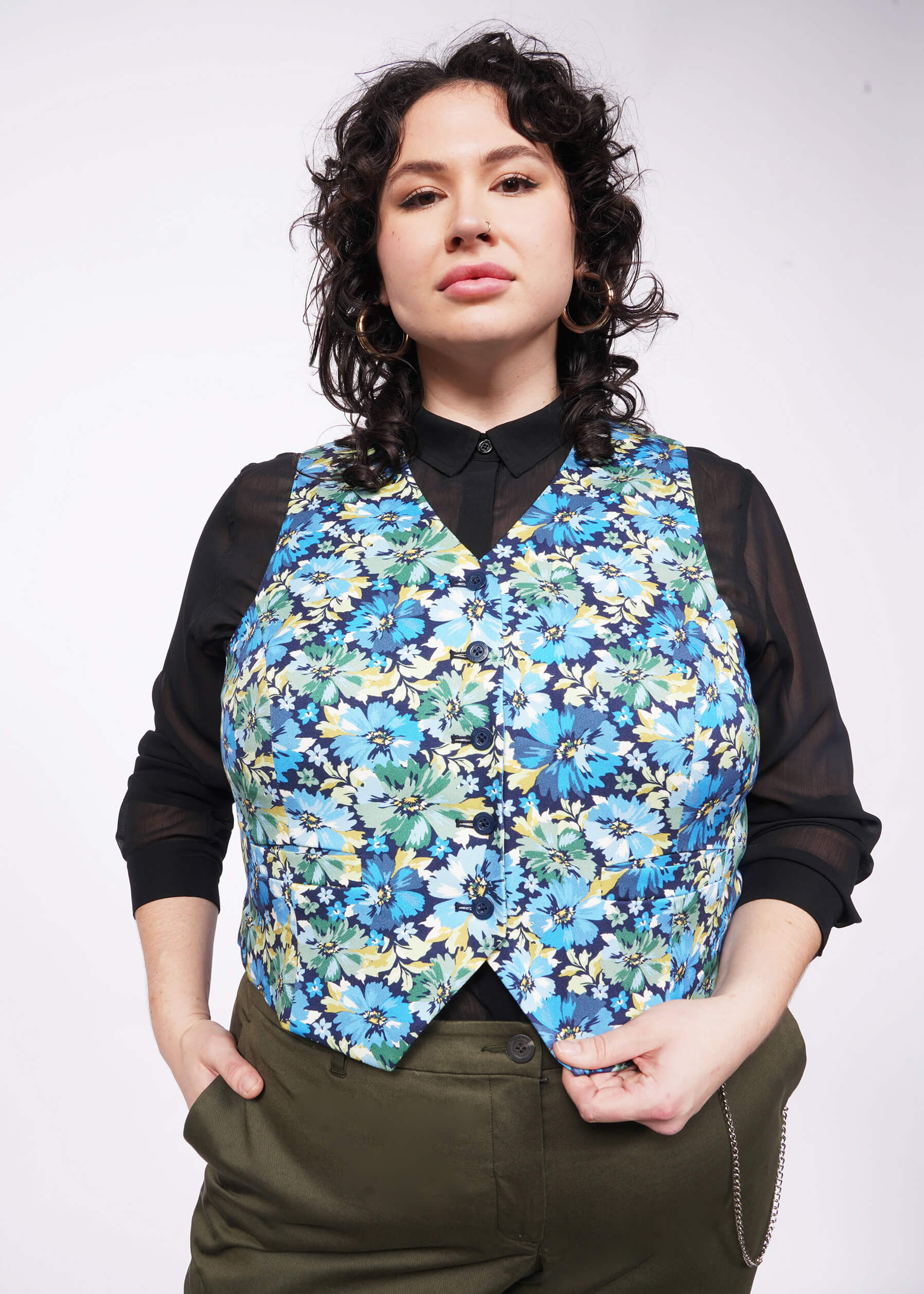 Person with black short curly hair wears a black sheer button up, The Empower Vest in Carnation Navy, which is a blue toned floral pattern, and green pants.