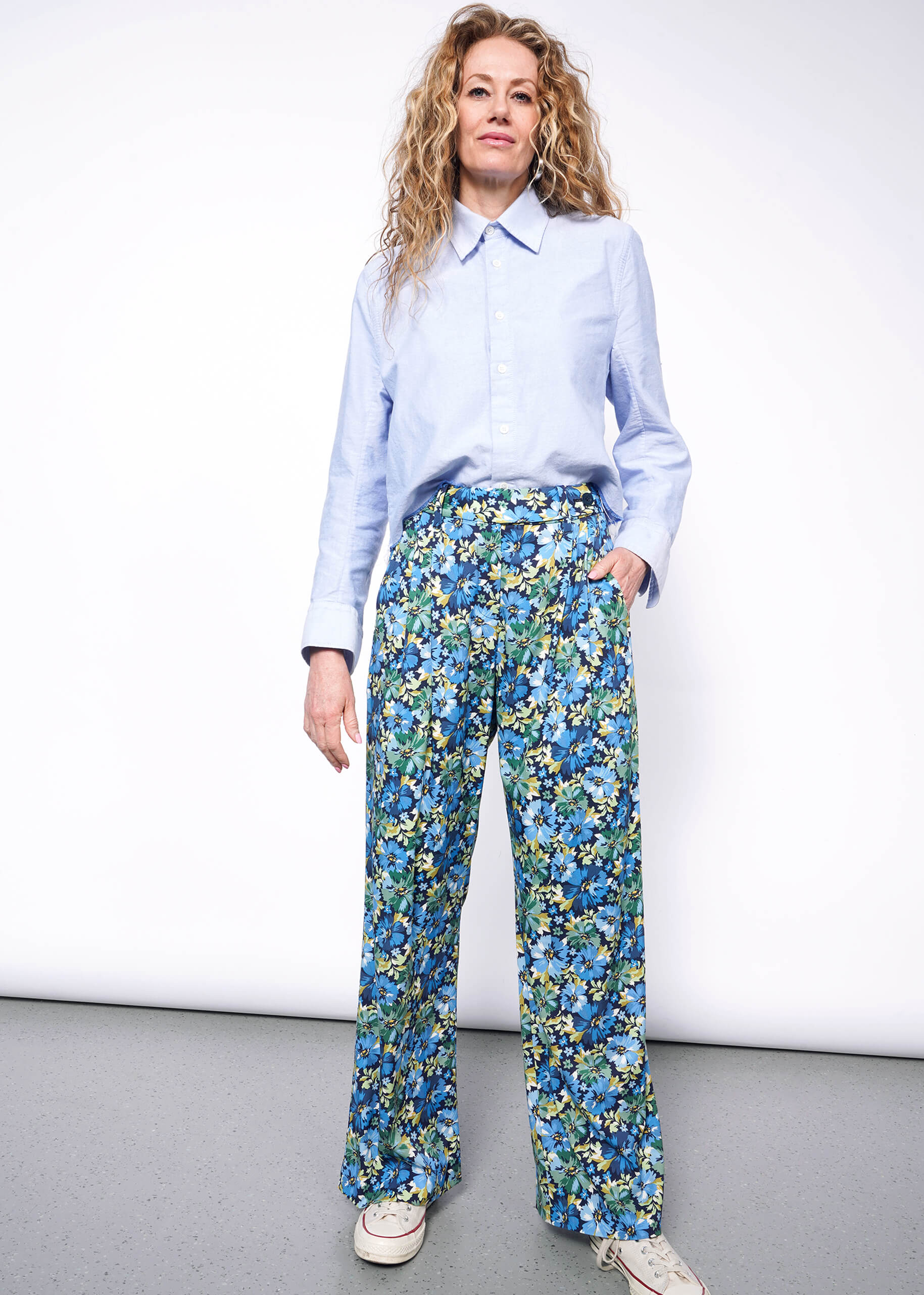 Person with wavy blonde hair stands looking at camera wearing a light blue button up and The Empower Wide Leg Pant in Carnation Navy, which is a blue toned floral pattern, with white Converse sneakers.