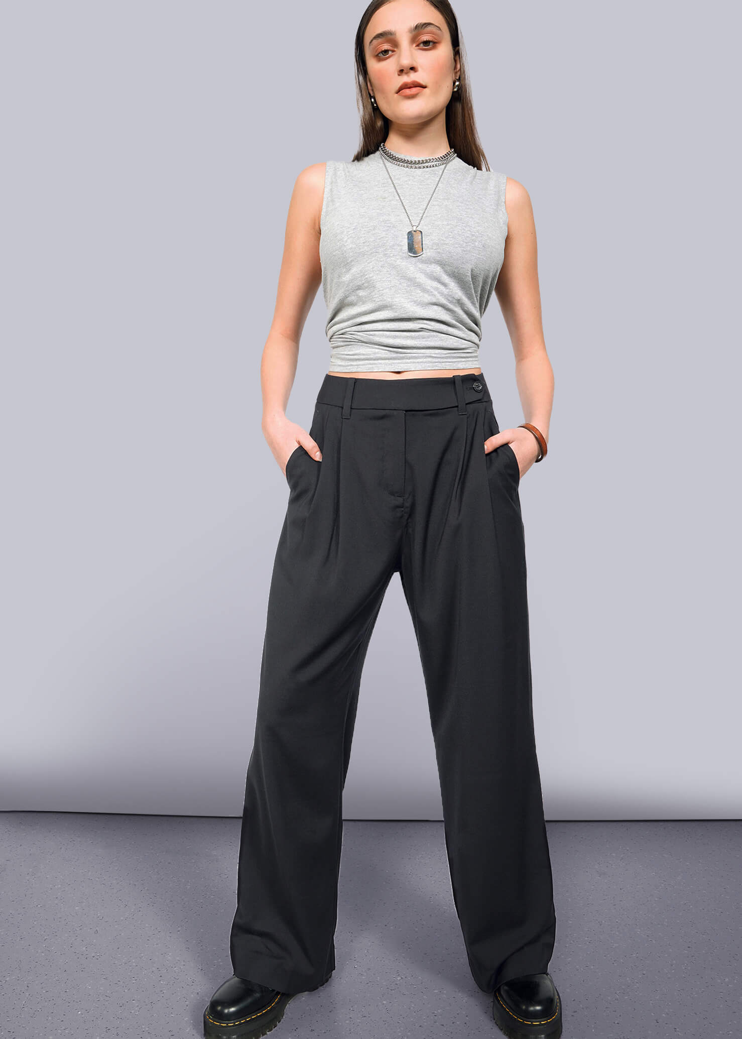 A person stands confidently against a white background, wearing The Empower Wide Leg Trouser with high waist and pleated front, paired with a gray sleeveless crop top, black shoes, layered necklaces, and hands in their pockets.