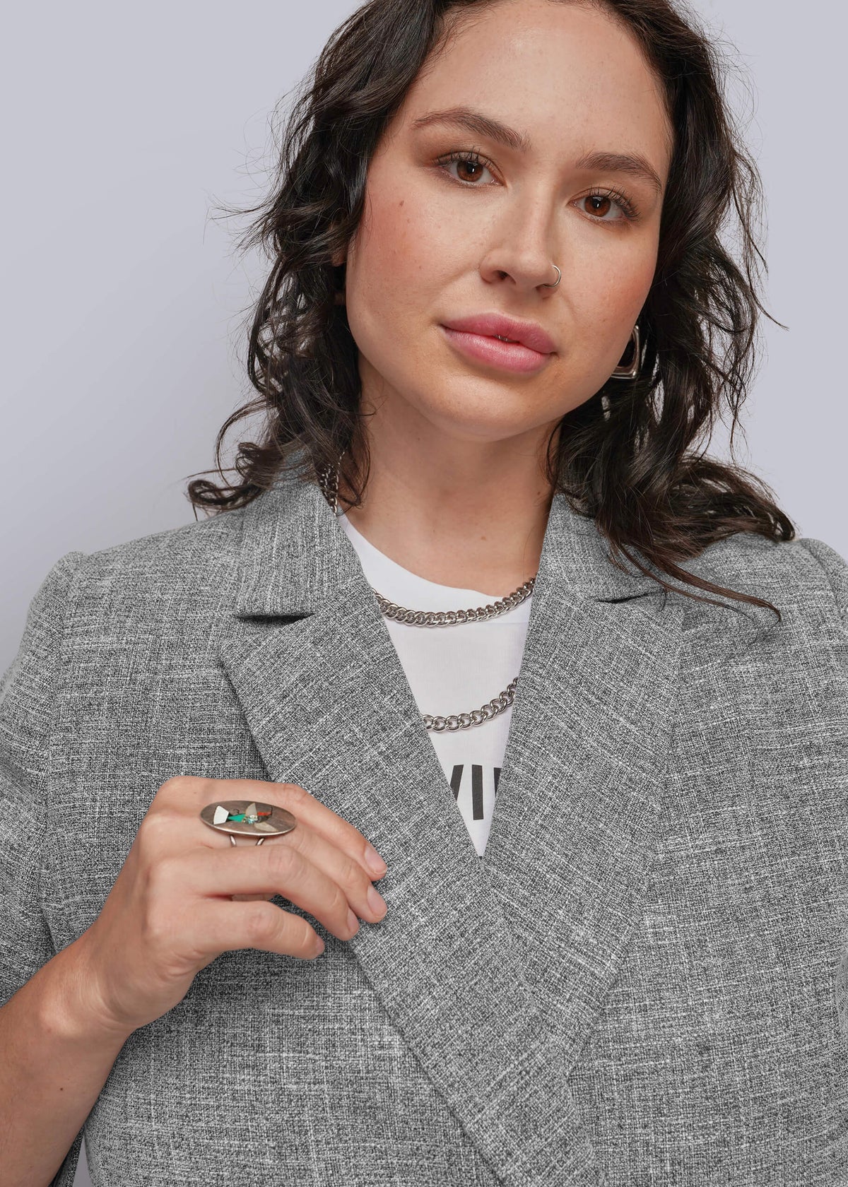1X=A close-up of a woman wearing a textured gray blazer over a white t-shirt, accessorized with silver chain necklaces and a green ring.