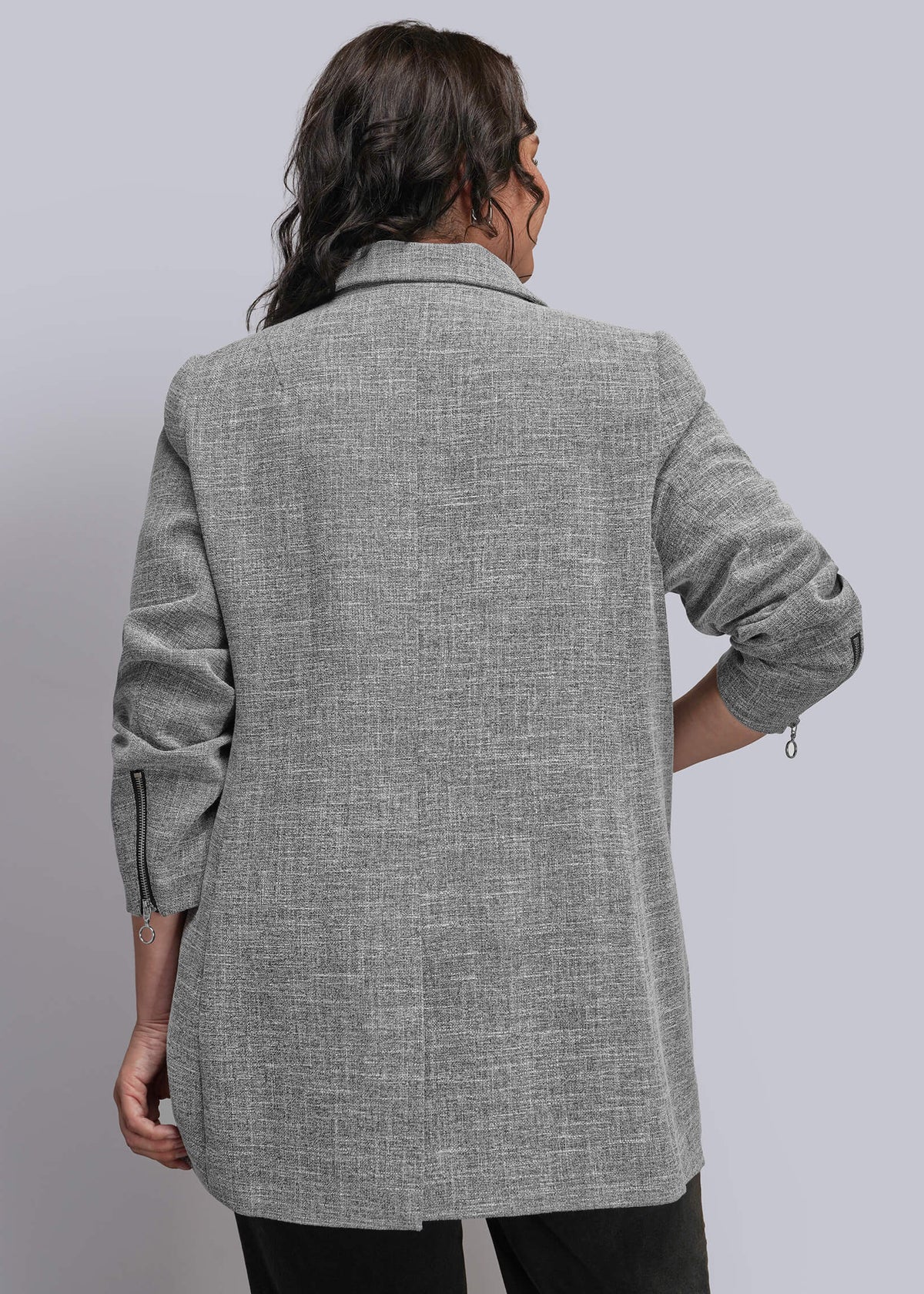 1X=A person shown from the back wearing a light gray textured blazer with rolled sleeves and black pants, photographed against a plain light gray studio background.