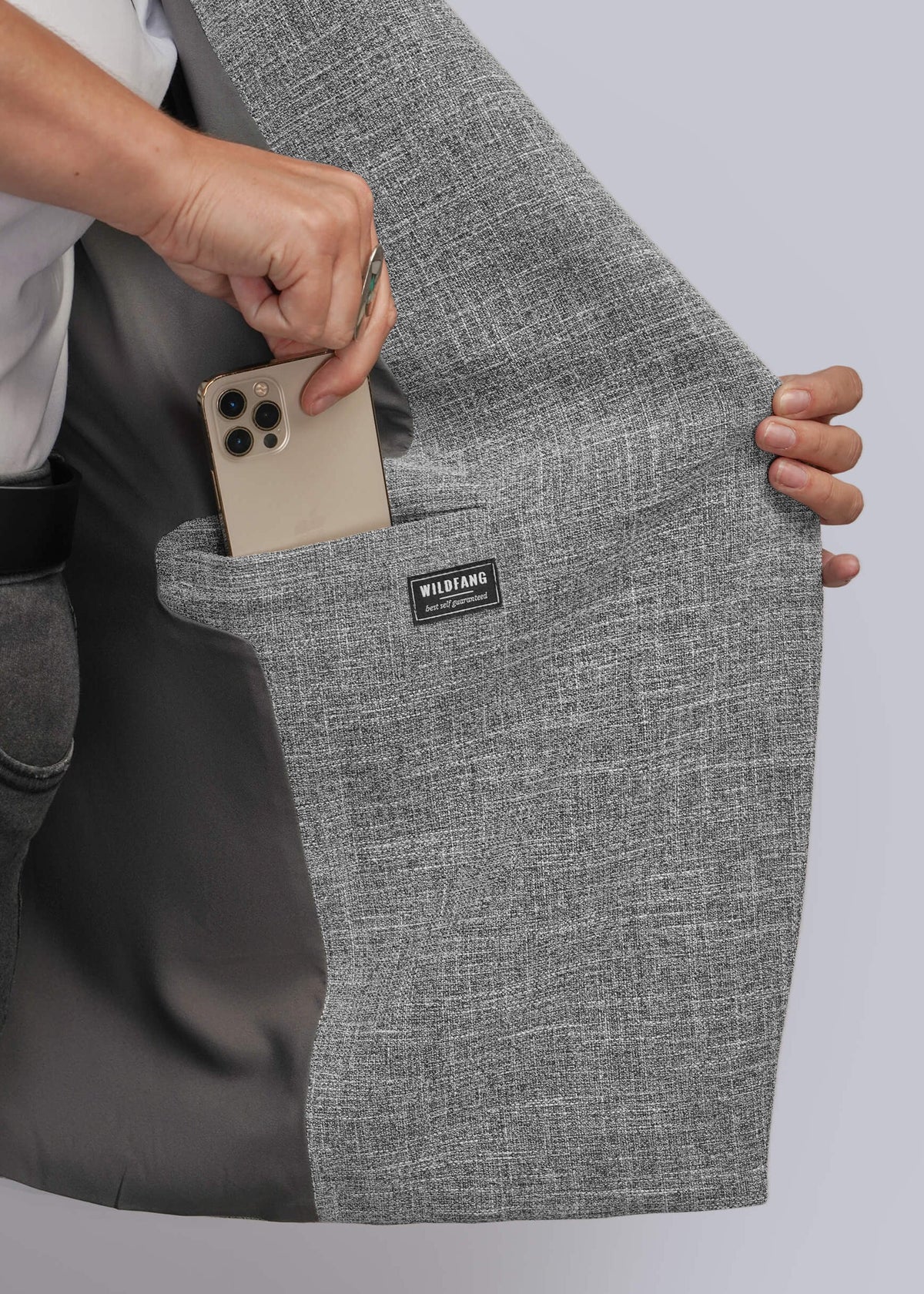 1X=Close-up of a person holding open a gray textured blazer to show a large interior pocket containing a smartphone. A small black brand label reading WILDFANG is visible on the pocket.